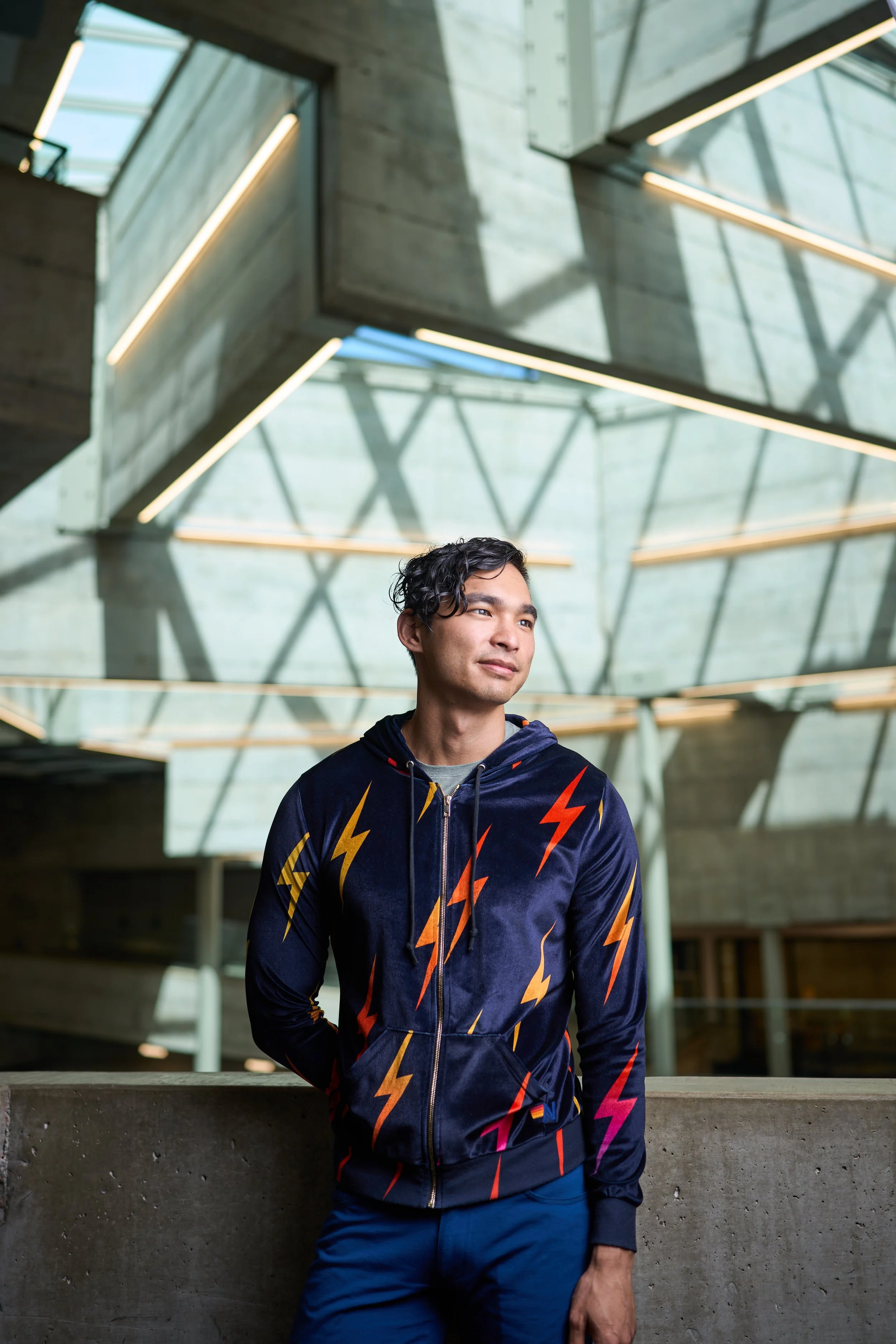A young man stands in a modern building with concrete walls and geometric glass windows, wearing a navy hoodie with orange and yellow lightning bolt patterns.
