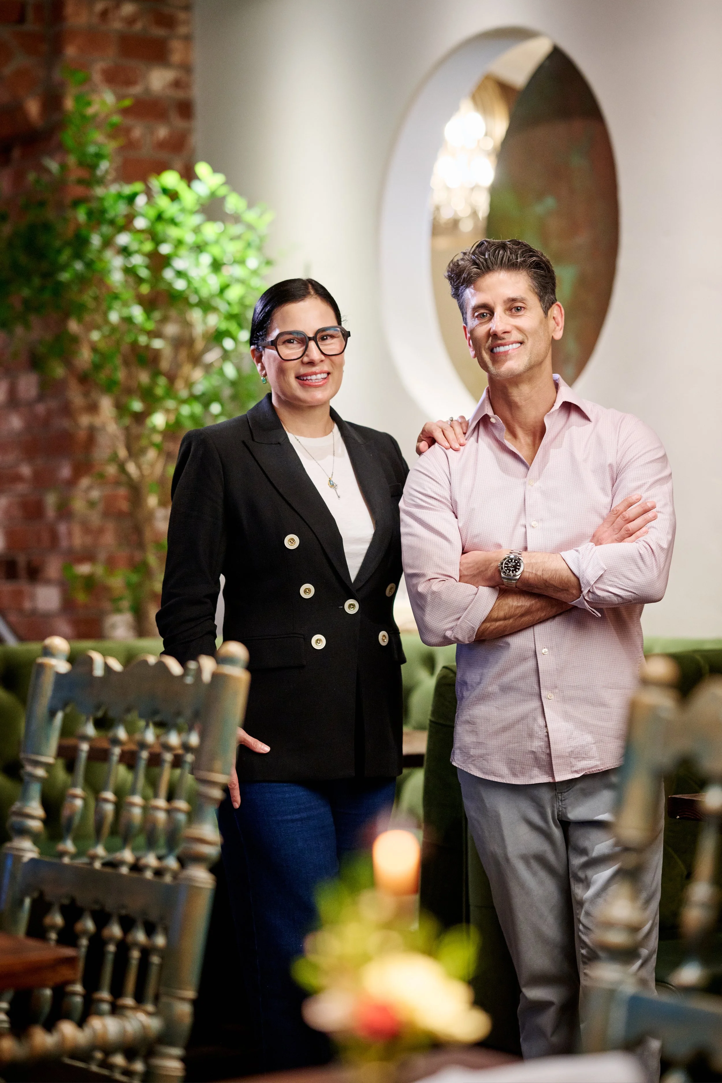 A woman with dark hair and glasses in a black blazer standing next to a smiling man with brown hair and a light pink shirt, both in a warmly lit indoor setting with a brick wall and green plant in the background.