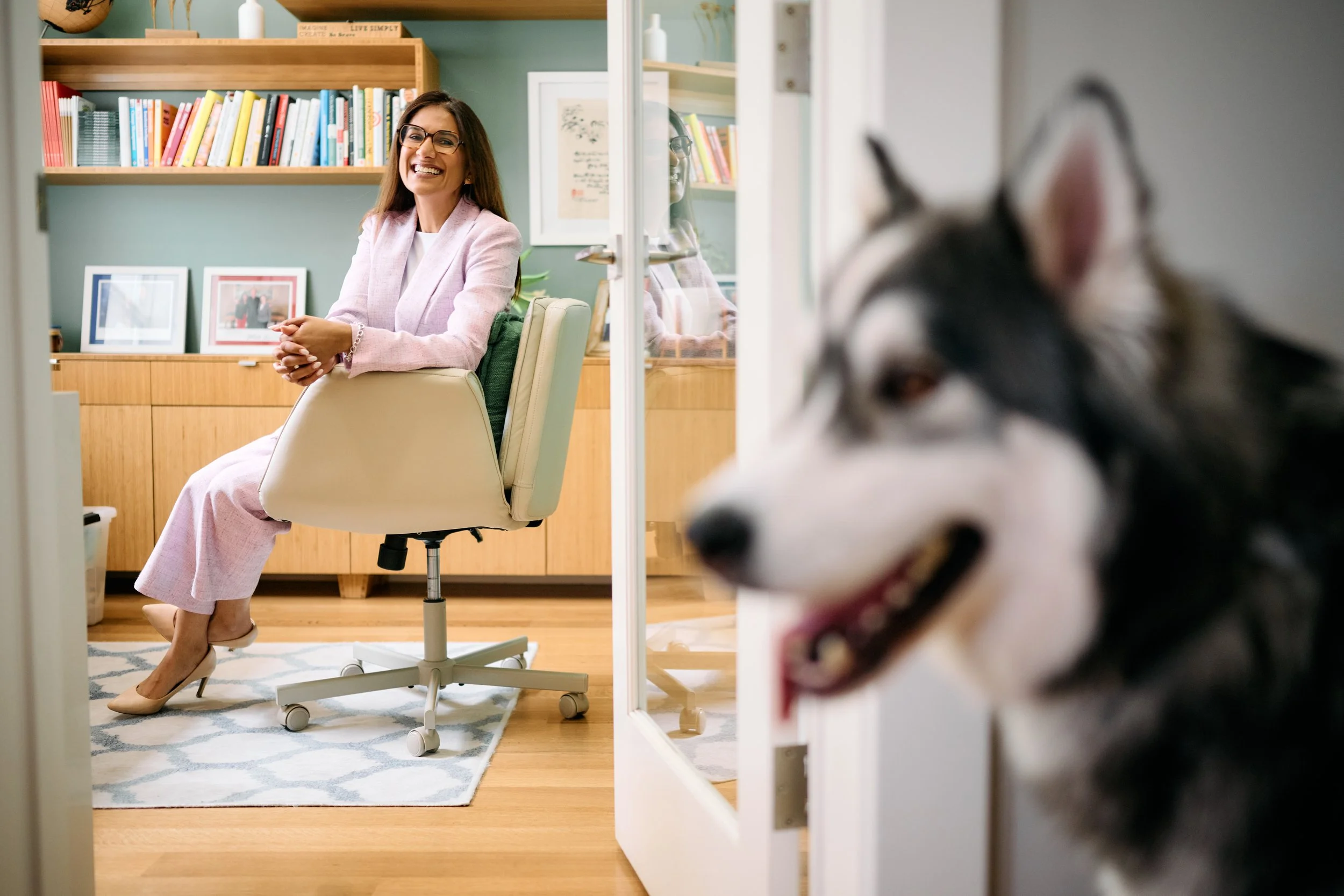 A woman with long brown hair, glasses, wearing a pink blazer and skirt, sits on a white office chair, smiling. She is in a room with a bookshelf, framed photos, and a plant. A dog is partially visible in the foreground, blurry, with its head turned t