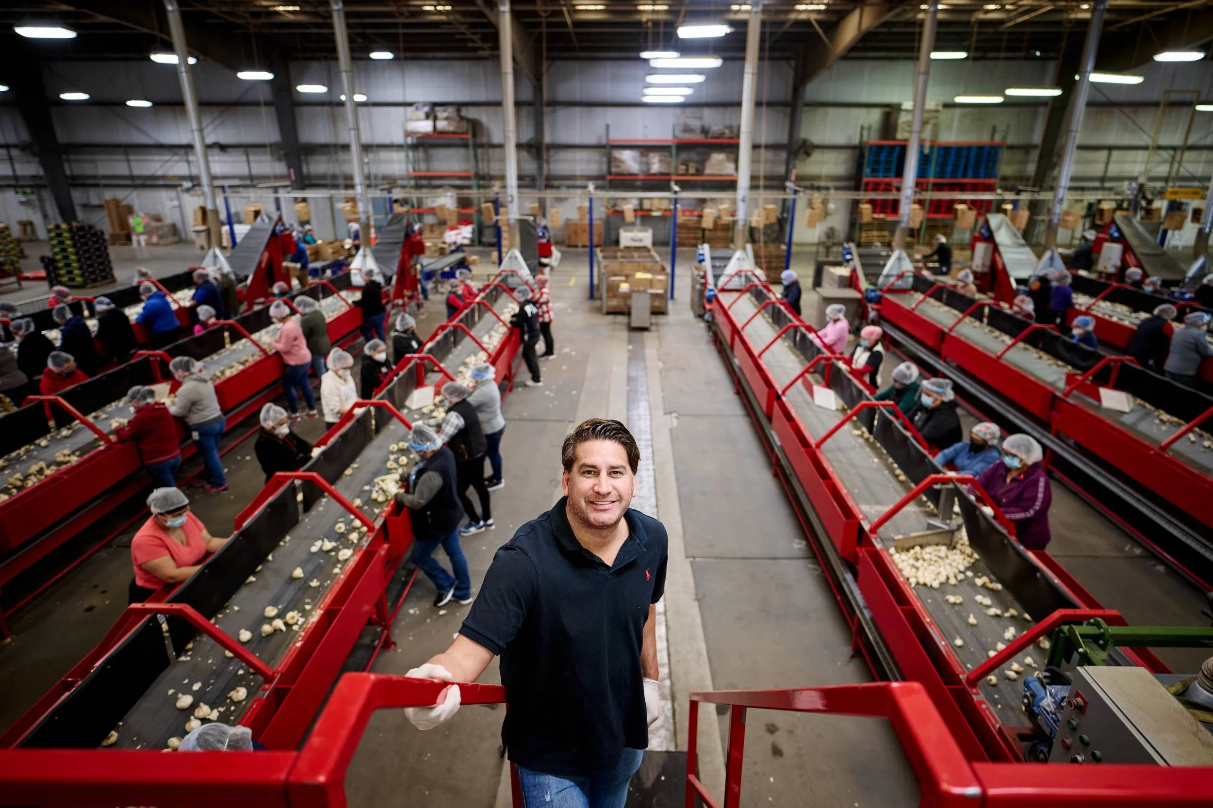 A factory worker standing in front of a conveyor belt filled with potatoes in a large industrial warehouse, with multiple workers wearing hair nets and masks working in the background.