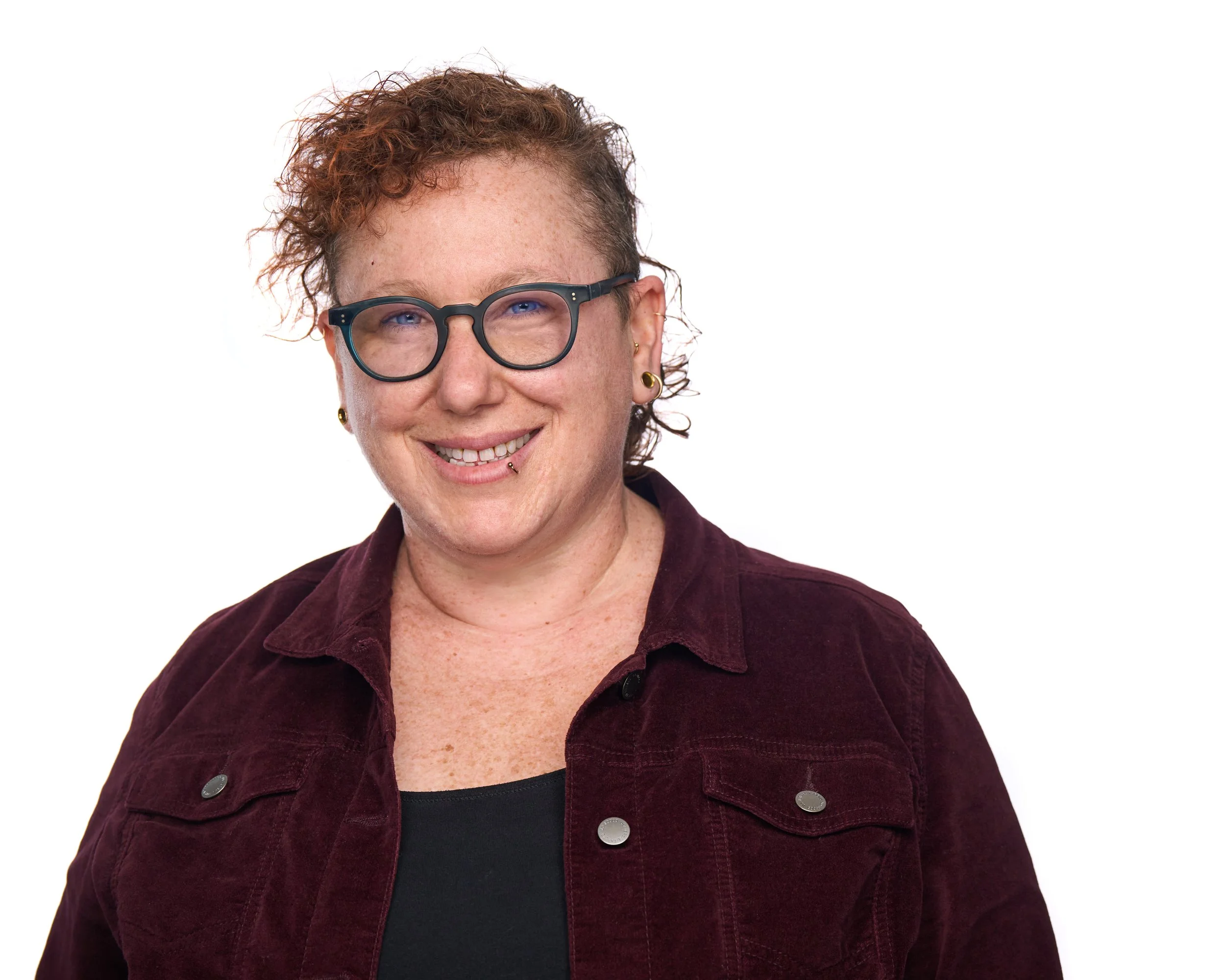 Portrait of a smiling person with short curly hair, glasses, and earrings, wearing a maroon jacket over a black top, against a white background.