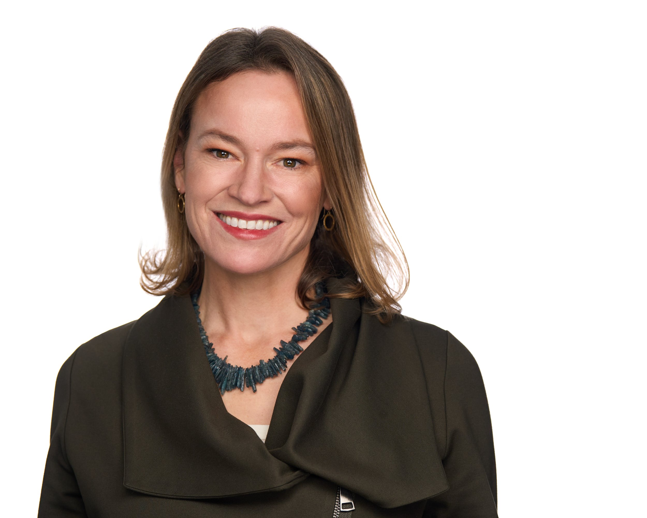 A woman with shoulder-length light brown hair smiling, wearing a dark blazer, a blue beaded necklace, and gold earrings, against a plain white background.