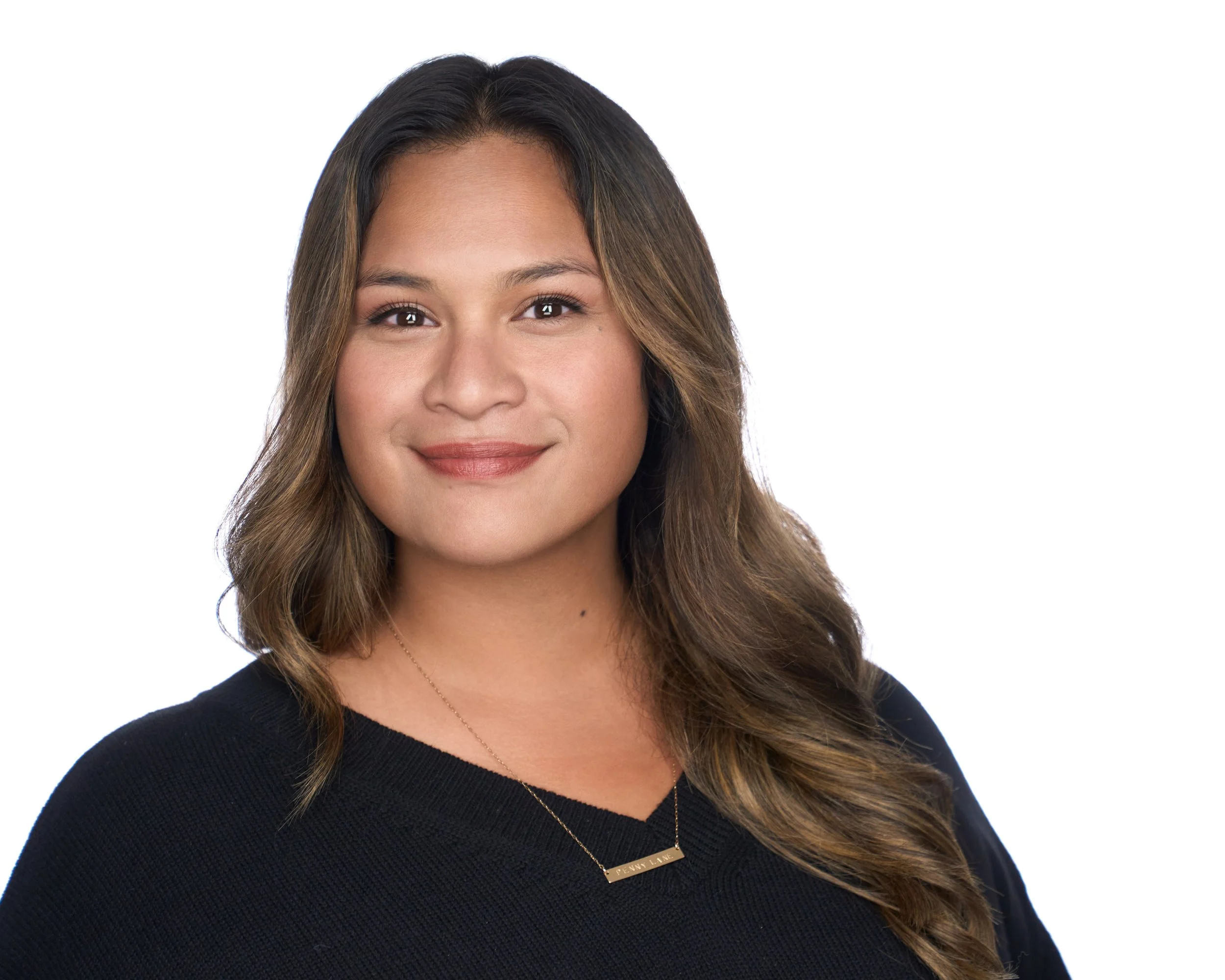 A woman with long wavy brown hair wearing a black top and a gold necklace smiling against a white background.