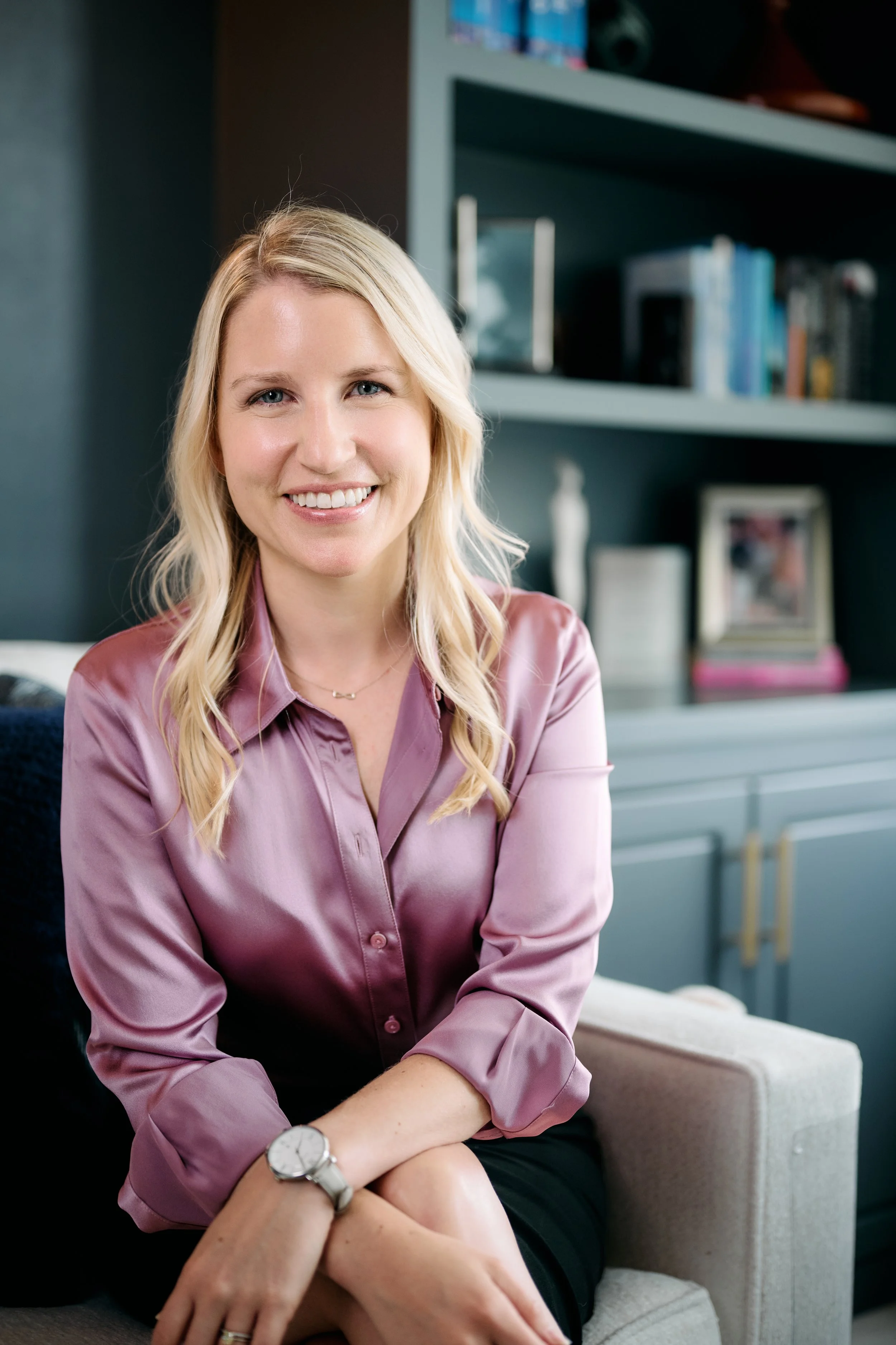 A smiling woman with blonde hair and blue eyes, wearing a pink satin blouse, sitting on a beige armchair in a room with a bookshelf and framed photos in the background.