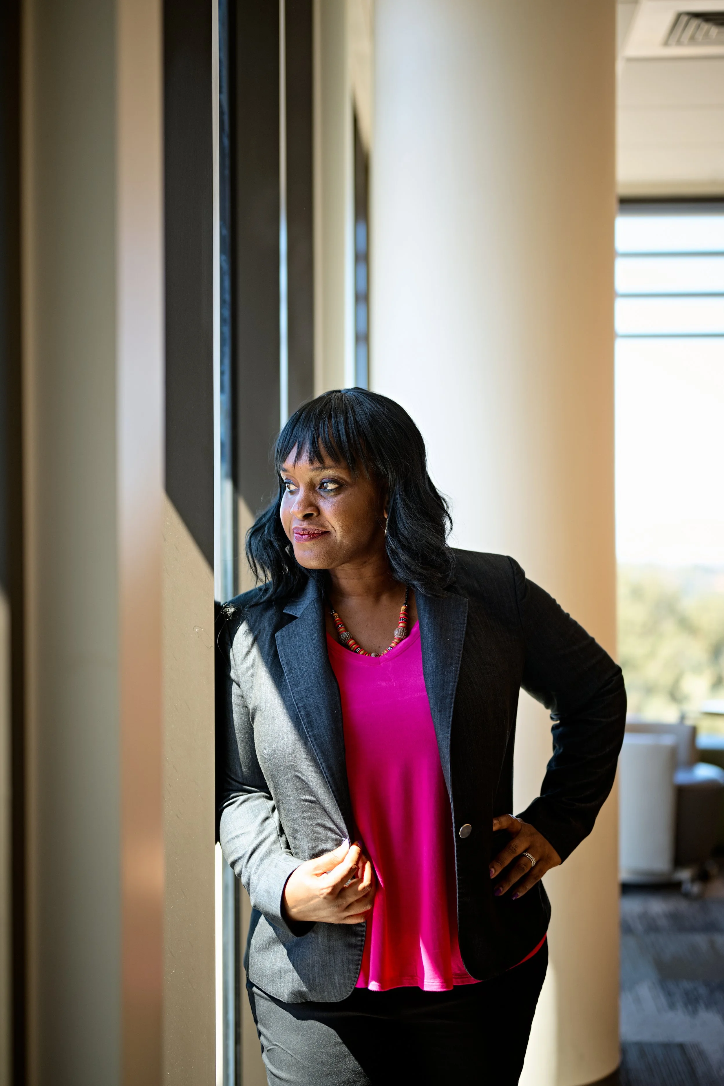 A woman in a business suit standing by a window in an office, looking outside with a thoughtful expression.