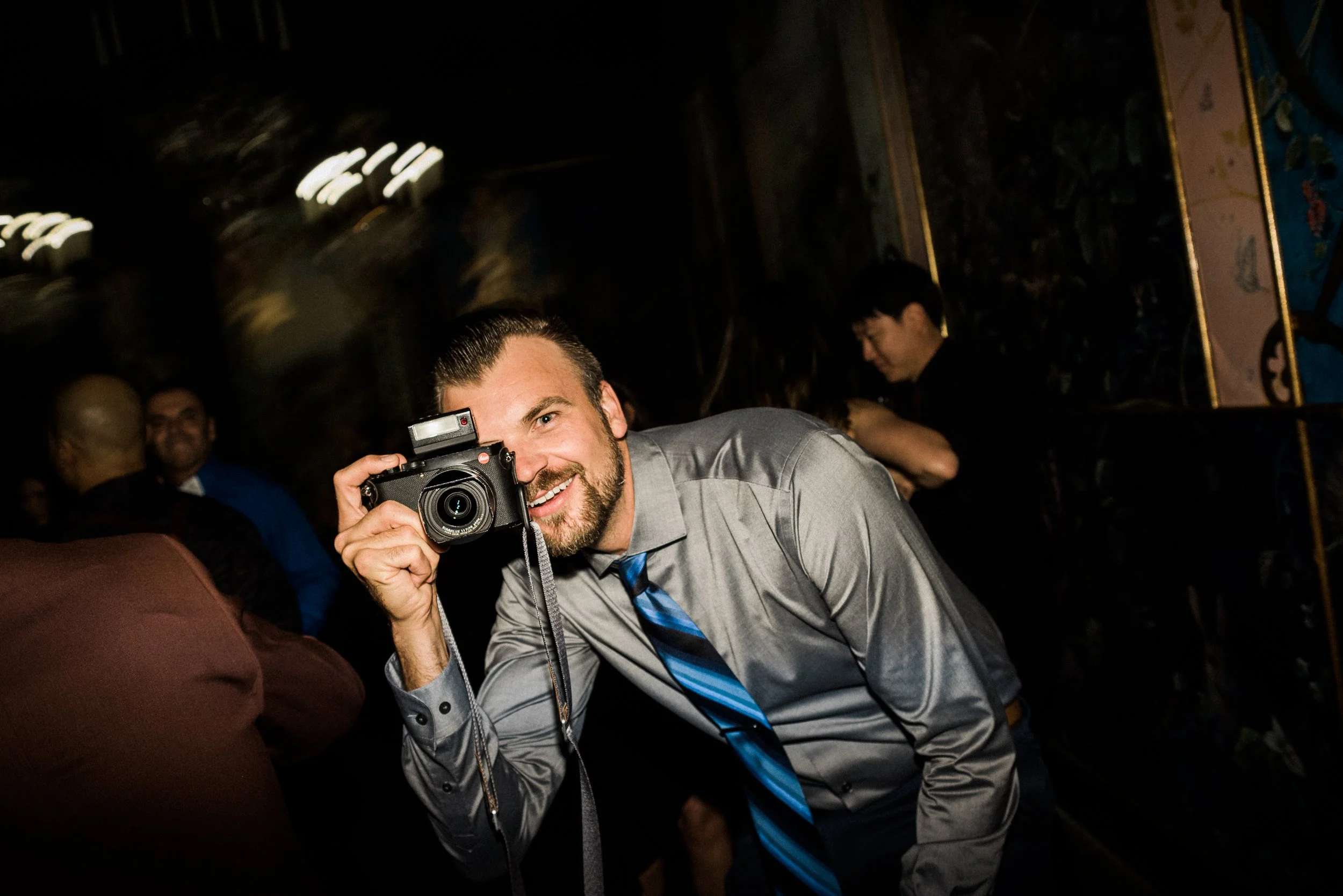A smiling man in a gray dress shirt and blue tie holding a camera to his face, looking directly at the lens, at a social gathering or event in a dimly lit room.