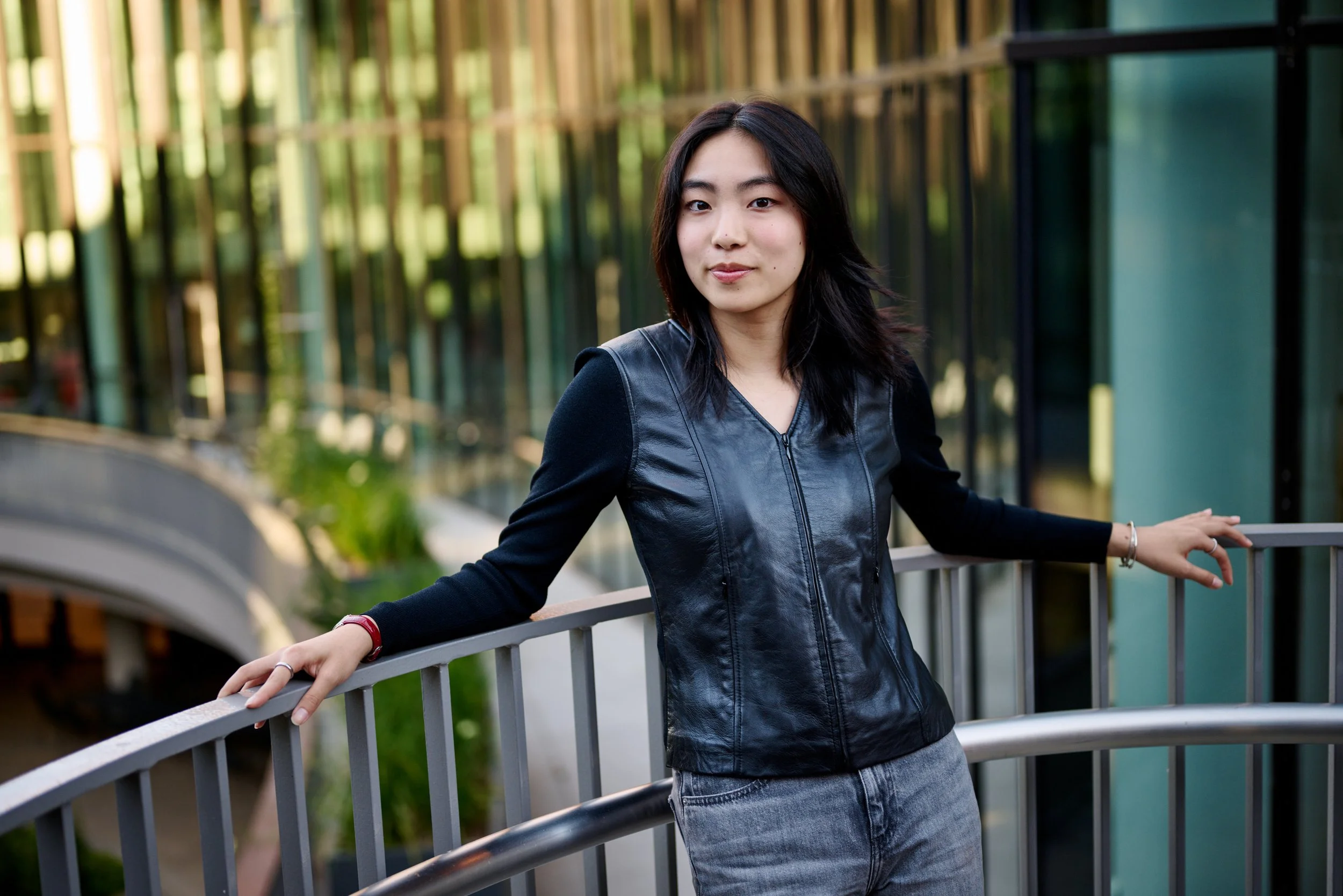 A young woman with shoulder-length black hair standing outdoors on a bridge, wearing a black leather vest, black long-sleeve shirt, and gray jeans, with one arm resting on the railing.