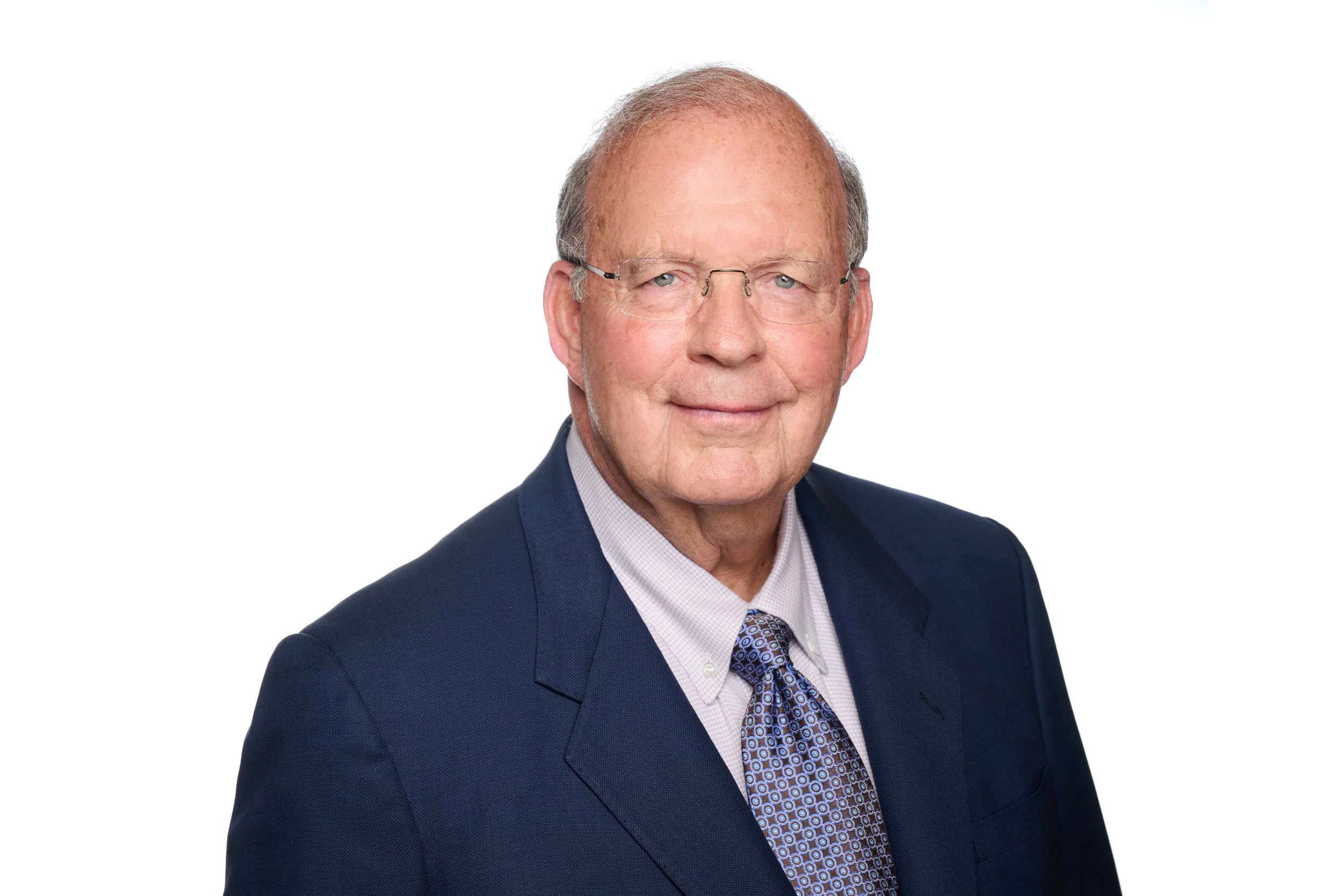 Portrait of an elderly man wearing glasses, a dark suit, a light shirt, and a patterned tie, smiling against a plain white background.