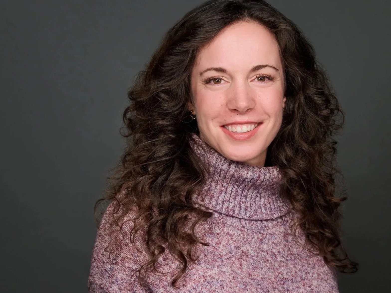 A woman with curly brown hair, wearing a maroon and purple turtleneck sweater, smiling against a dark background.
