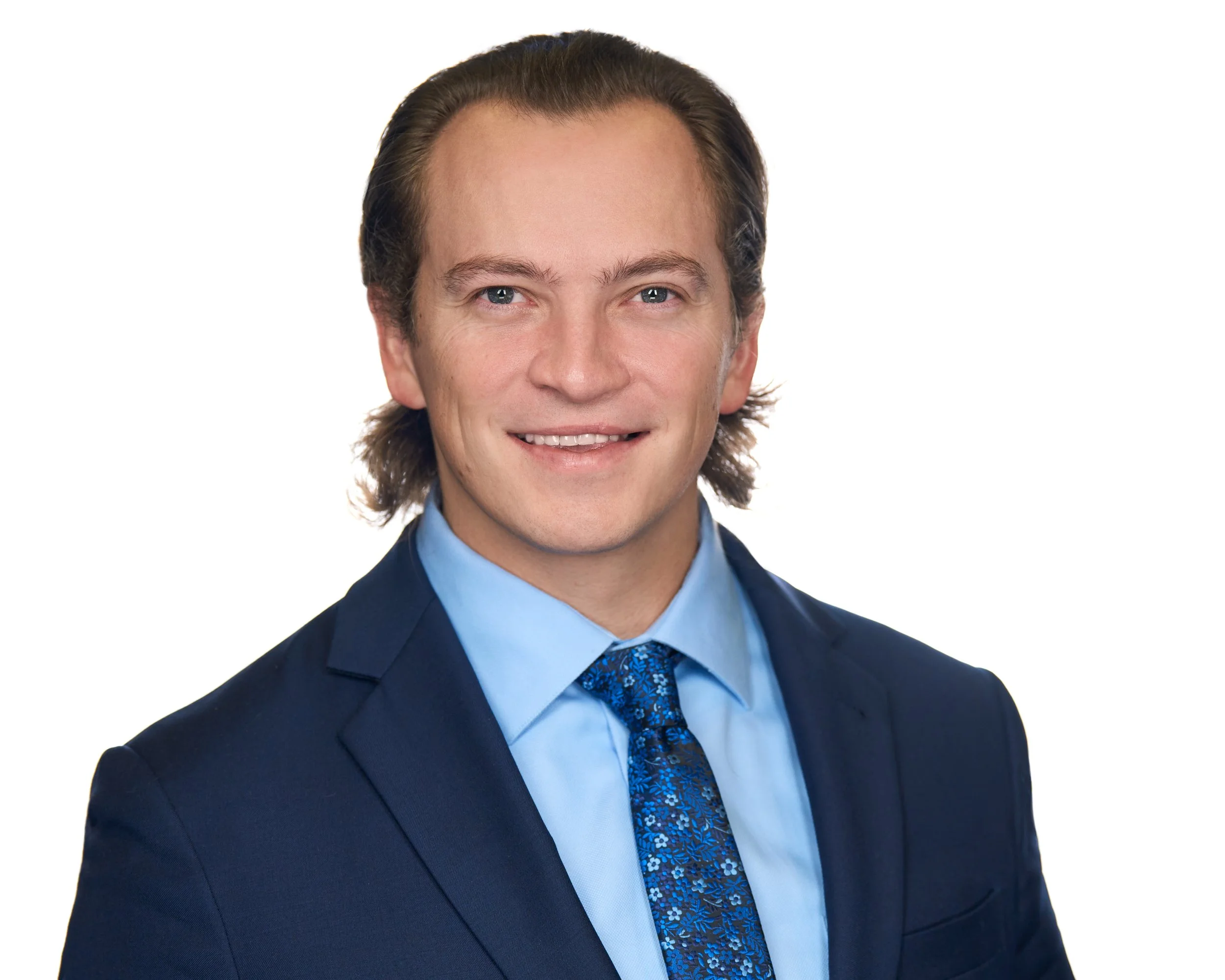 Professional headshot of a young man in a dark suit with light blue dress shirt and patterned tie, smiling against a white background.