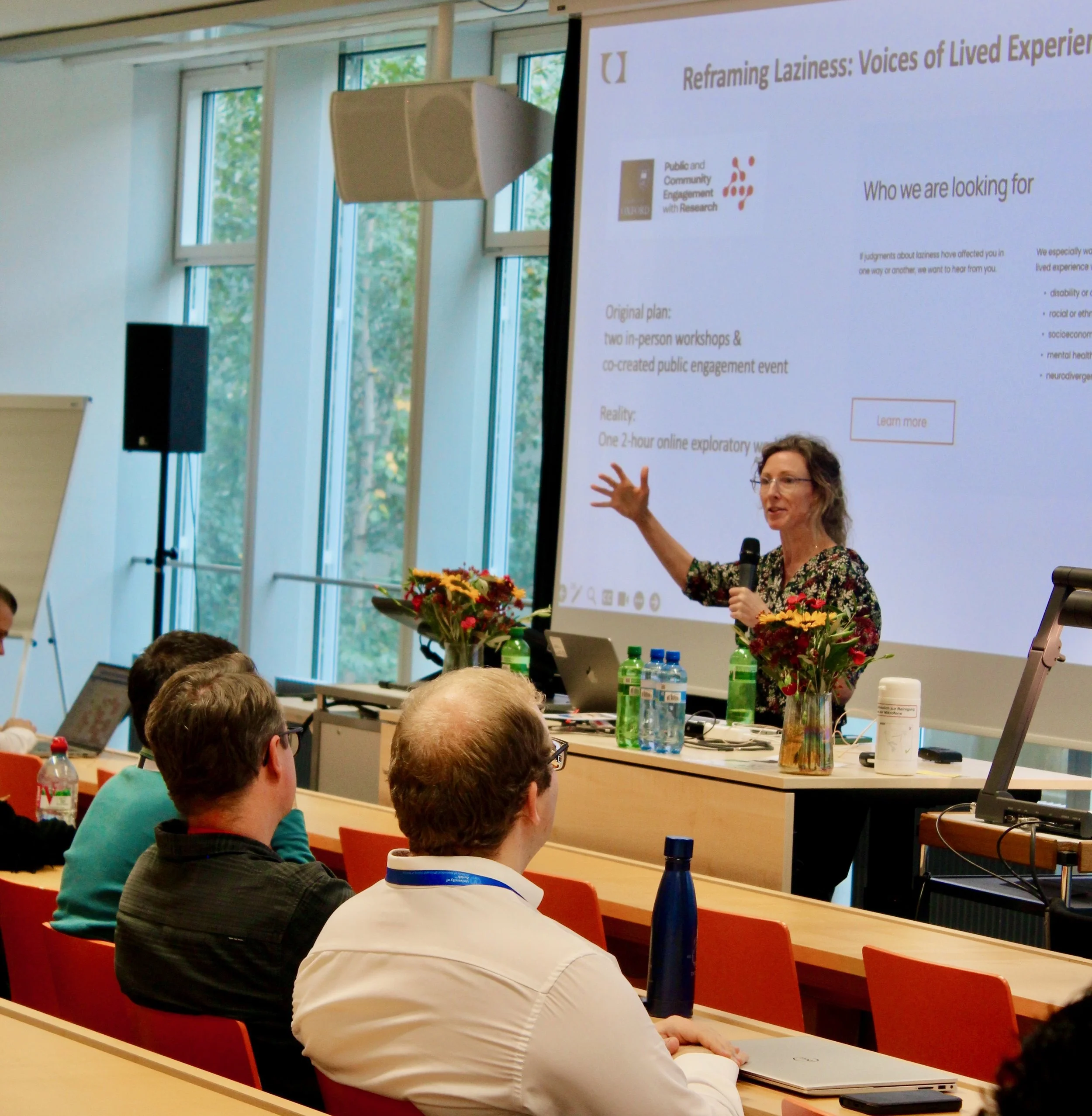 Professor Katrien Devolder, lead of Project Lazy, speaks to an audience at the front of a lecture theatre. A large screen behind her displays the presentation title, Reframing Laziness: Voices of Lived Experiences.