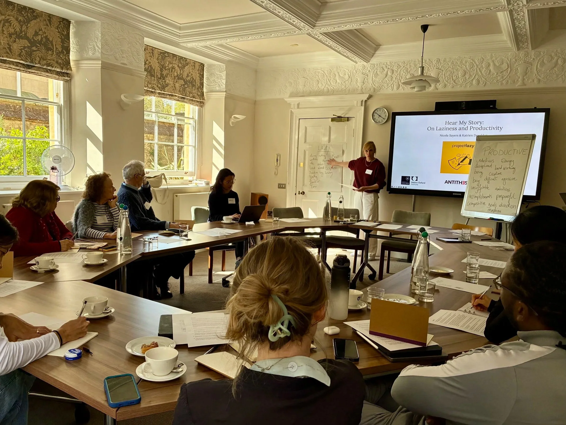 Writer Nicola Sayers leads the workshop from the front of a bright meeting room. Attendees sit around U-shaped tables, listening and taking notes. Flip charts in front of the room show words associated with 'Productive' and 'Lazy'.