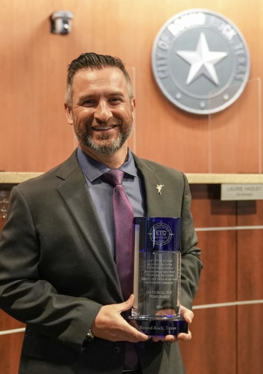 Round Rock Mayor Craig Morgan smiling and holding the ETC Institute National Top Performer award trophy in the Round Rock City Council chambers, with the City of Round Rock seal visible on the wall behind him.