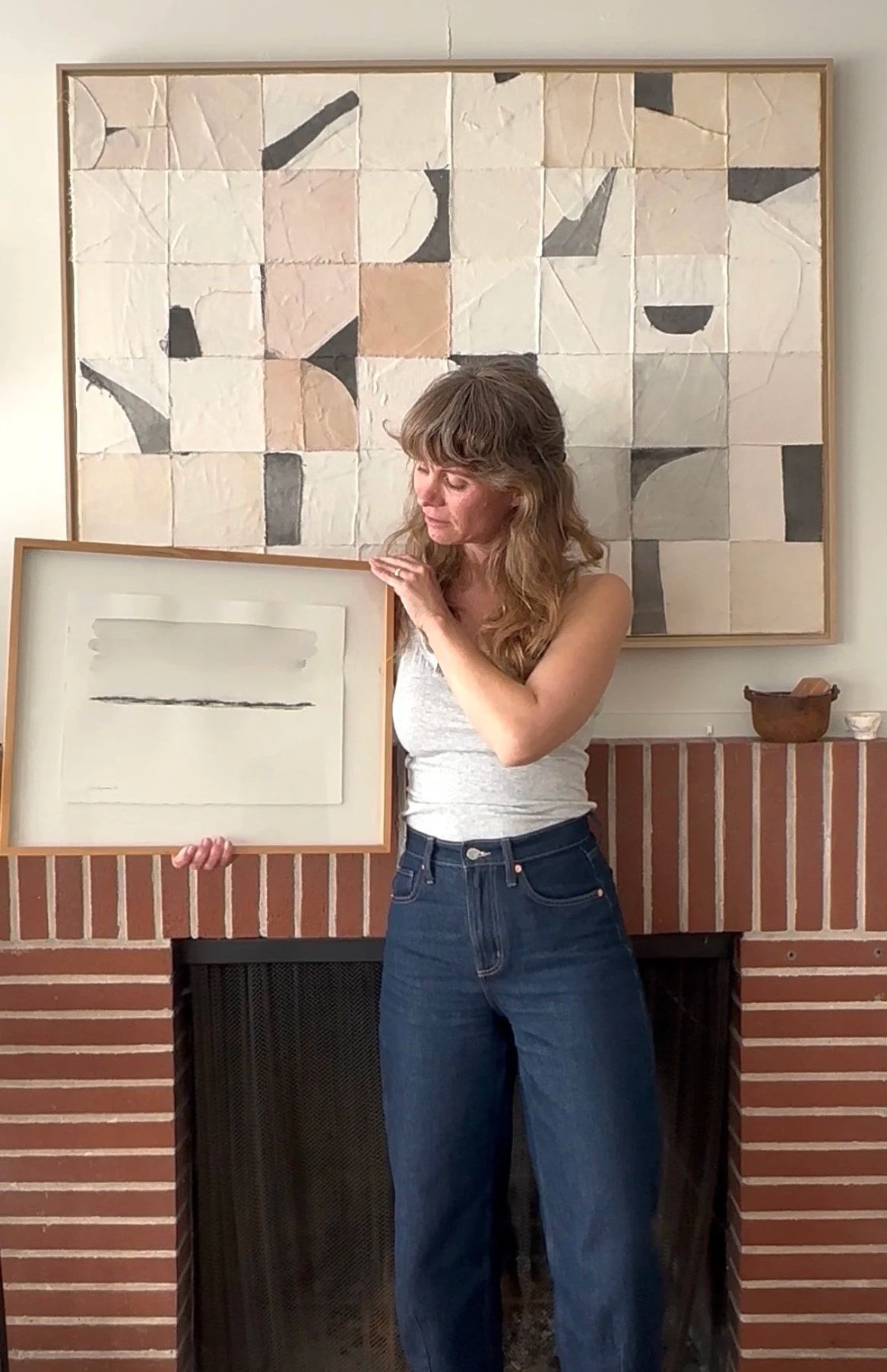 A woman with wavy light brown hair wearing a gray tank top and blue jeans stands in front of a brick fireplace. She is holding a framed piece of artwork with a white background and a single black line, and is looking down at it. A large abstract painting with geometric shapes and neutral colors hangs on the wall behind her.