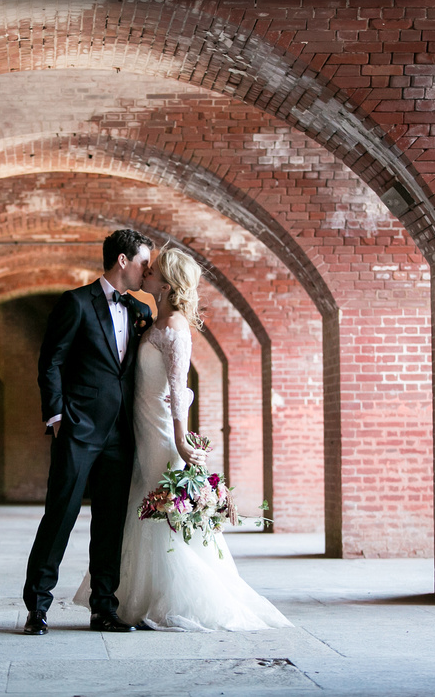 Bride and groom kissing under brick arches in an elegant setting, bride holding a bouquet.