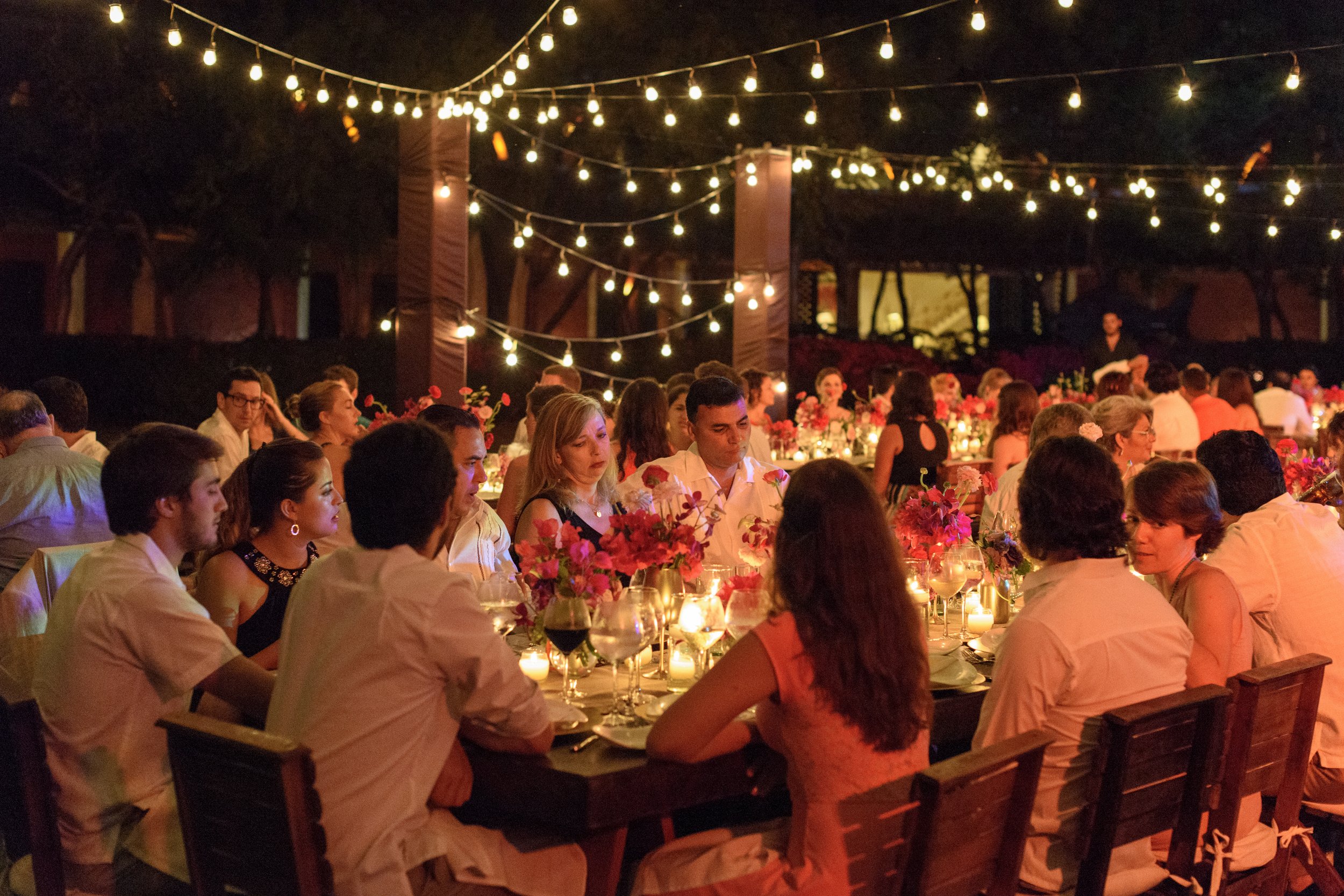 People dining at an outdoor evening event with string lights and floral decorations.