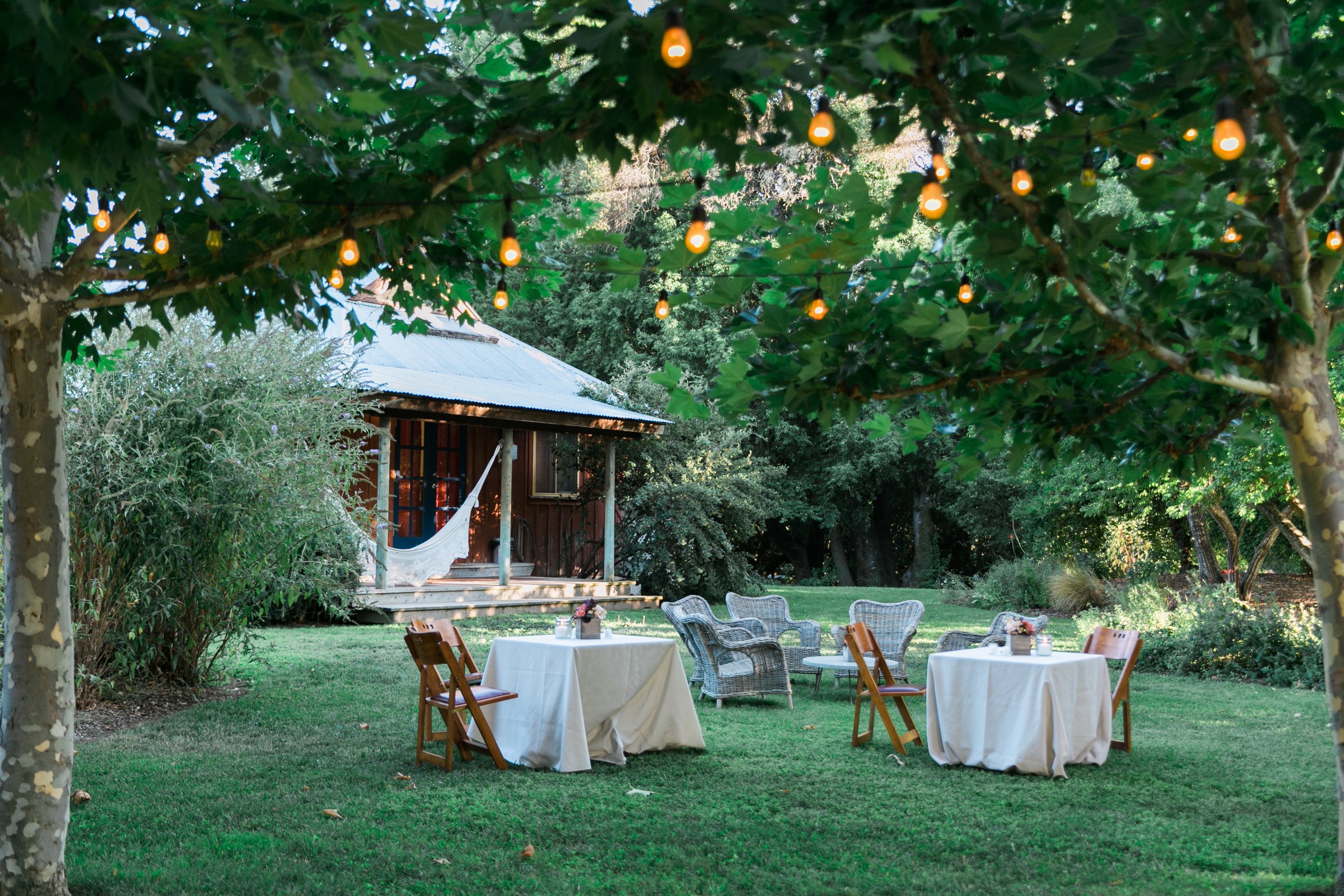 Outdoor scene with decorated tables and chairs on a lawn, surrounded by greenery and trees with hanging string lights, near a rustic cabin with a hammock on the porch. Boonville hotel wedding.