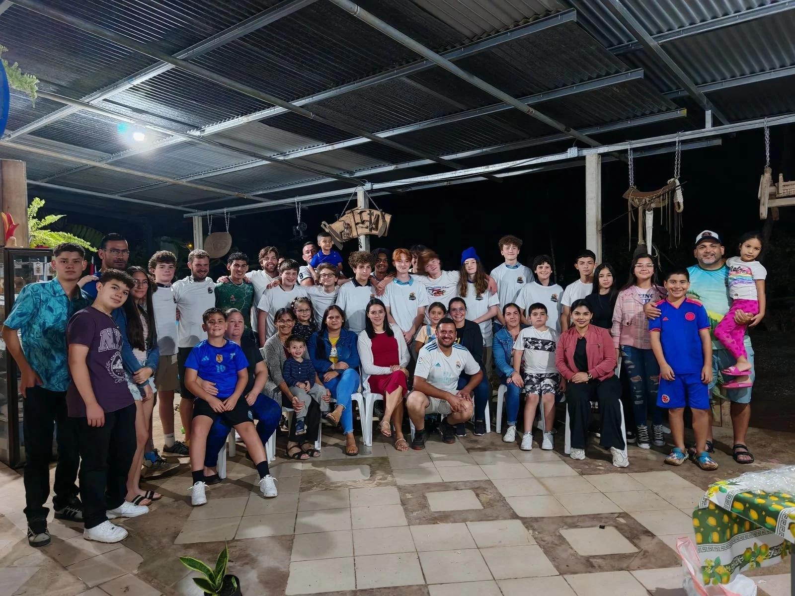 Grupo grande de personas, incluyendo niños y adultos, posando para una foto en un espacio cerrado con techo de lámina, decorado con objetos culturales y plantas.