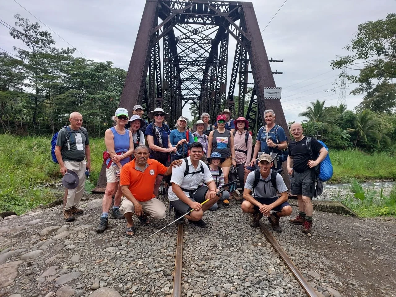 Grupo de personas haciendo senderismo frente a una estructura de puente de hierro en medio de vegetación tropical.