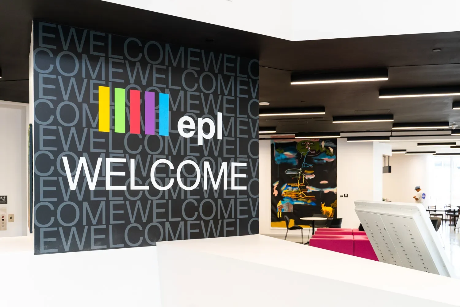 Interior of a modern building with a large wall display that reads "Welcome" and "epic" with colorful bars above the text. There are artworks, seating, and a person visible in the background.