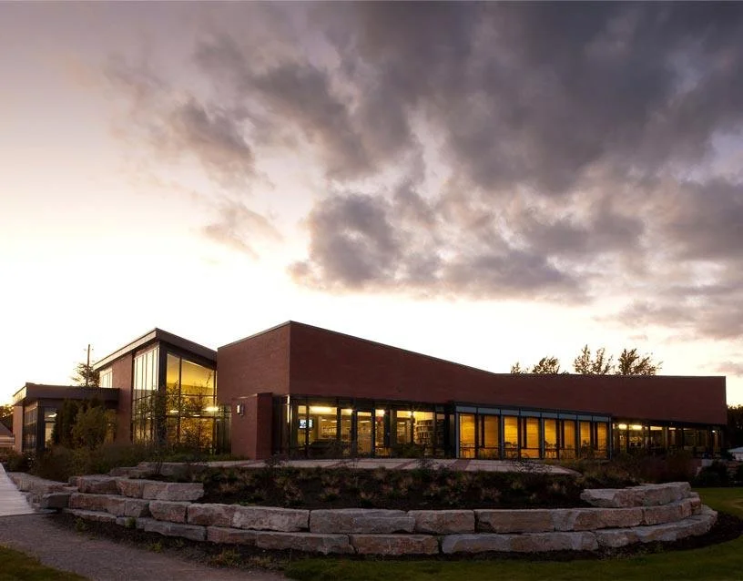 Modern building with large glass windows, illuminated from within, situated on a landscaped area with stone edging, against a cloudy evening sky.