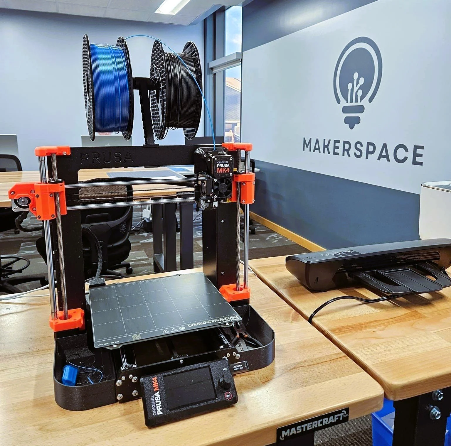 A 3D printer with filament spools on top at a makerspace, with a logo of a lightbulb with circuits on the wall in the background.