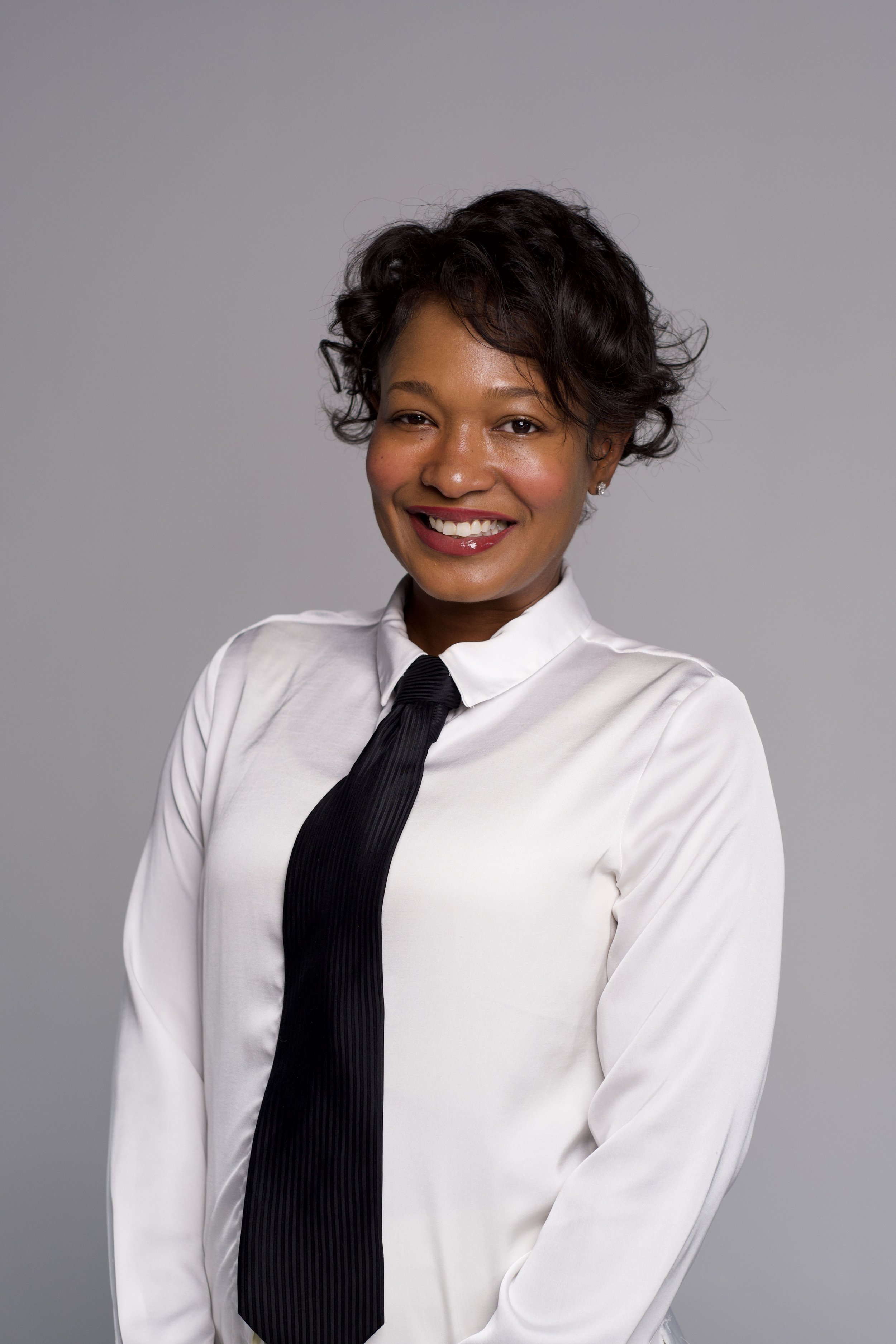 Portrait of a woman with long black hair wearing a black and white embellished blazer with pearl accents, posing against a dark background.