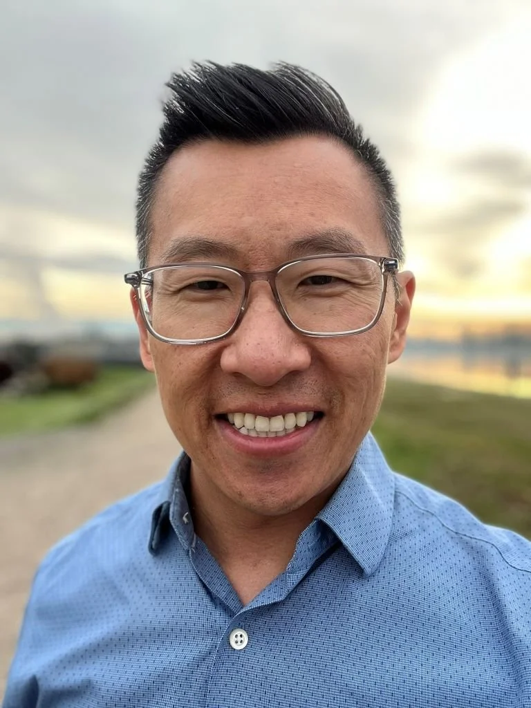 A smiling man with glasses taking a selfie outdoors with trees and sky in the background.