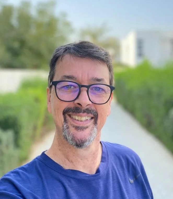 A smiling man with glasses and a goatee, wearing a blue shirt, outdoors on a sunny day with greenery and a pathway in the background.