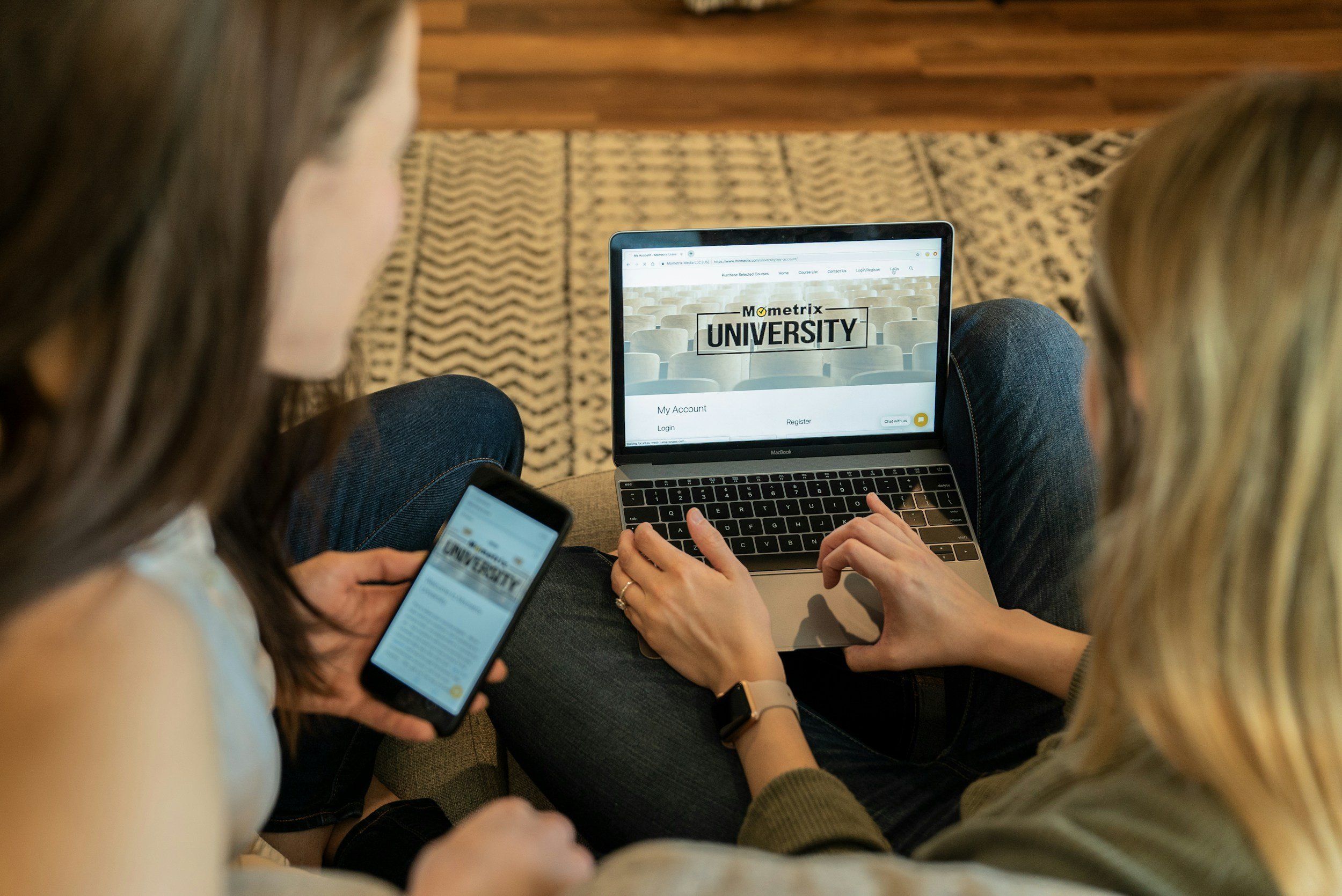 Two women sitting on a couch, one using a smartphone and the other using a laptop, both viewing the Mometrix University website.