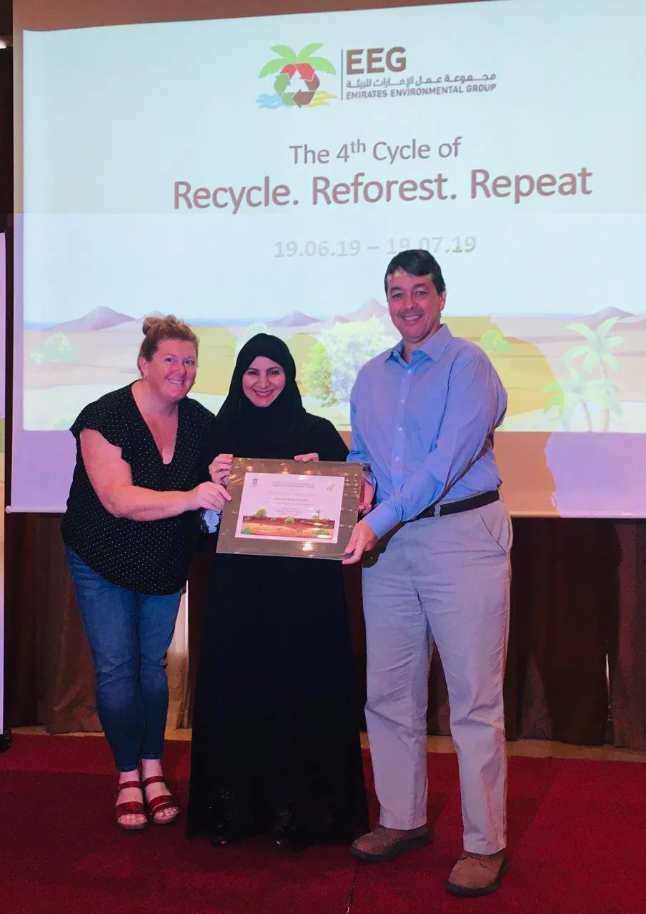 Three people, two women and one man, are standing together in front of a projector screen during an award ceremony. The woman in the middle is holding a framed certificate. The screen behind them displays the logo of Emirates Environmental Group and the text 'The 4th Cycle of Recycle. Reforest. Repeat,' along with the dates 19.06.19 to 19.07.19.