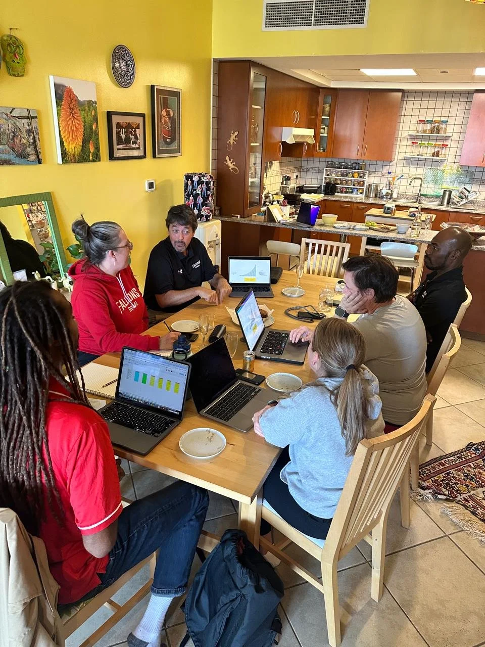 A group of six people sitting around a wooden dining table engaged in a meeting, with laptops, notebooks, and plates on the table, in a kitchen-adjacent dining area.