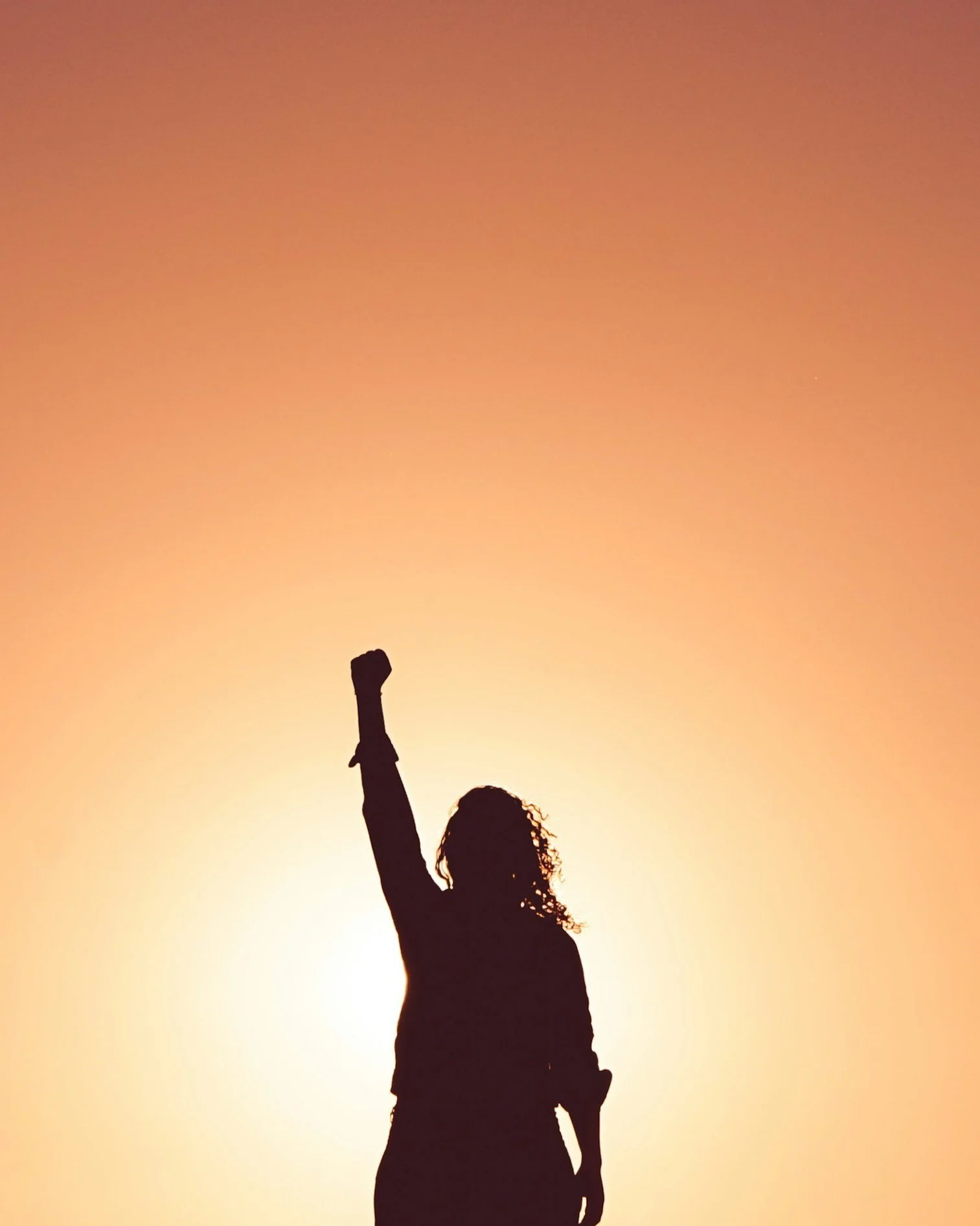 Silhouette of a woman raising her fist in the air with the sun setting behind her, creating an orange sky.