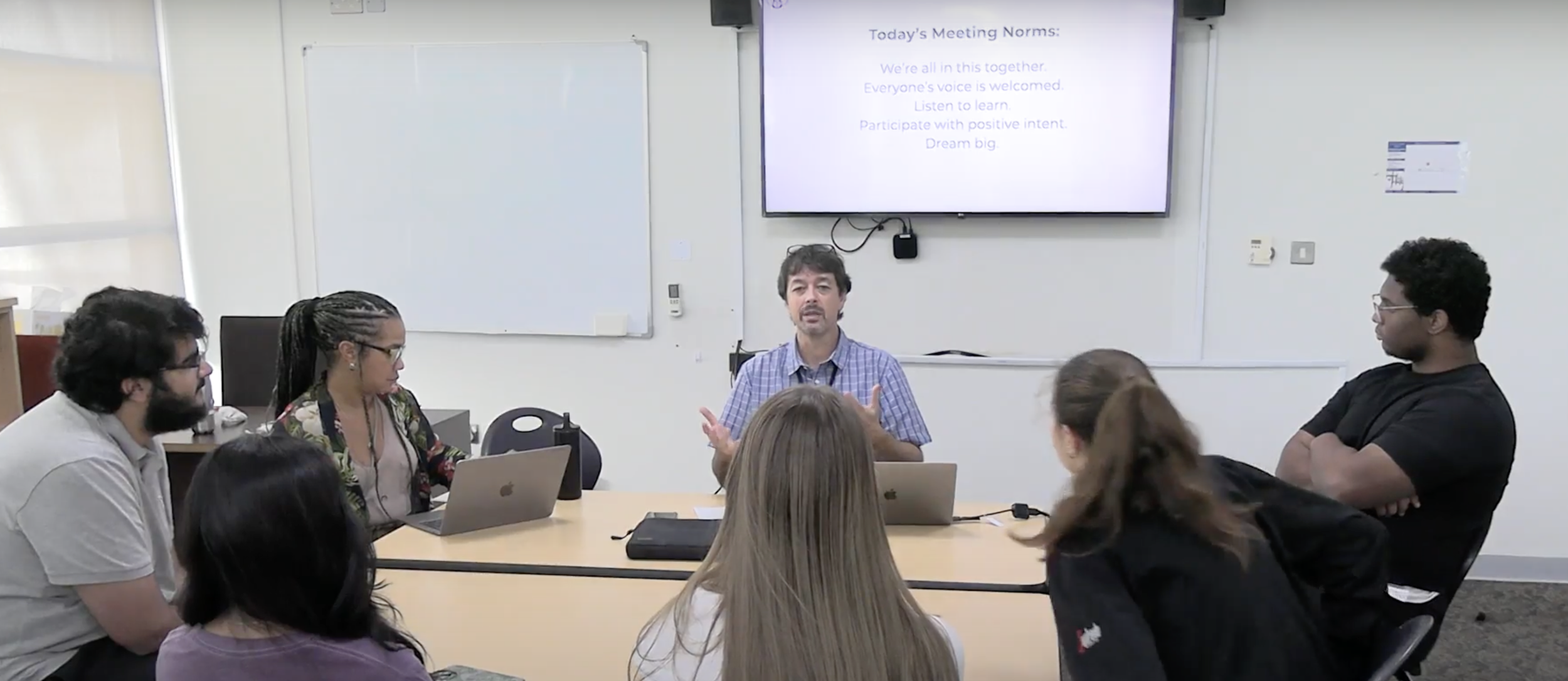 A group of people seated around a conference table in a meeting room, listening to a man speaking at the head of the table. There is a large screen behind him with the 'Today's Meeting Norms' displayed. The room has a whiteboard on the wall.