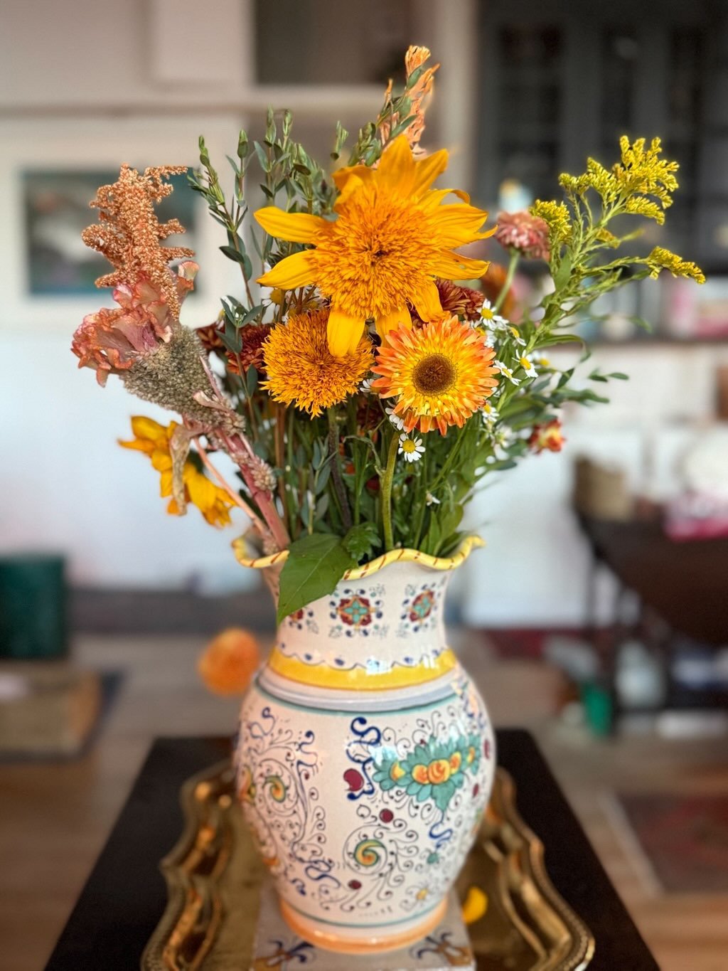 A decorative ceramic vase with colorful floral patterns contains a bouquet of vibrant yellow and orange flowers, placed on an ornate tray on a wooden table in a cozy indoor setting.