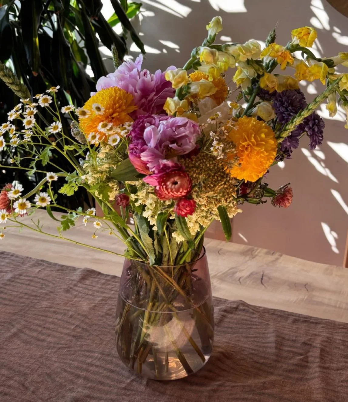 A glass vase with a colorful bouquet of assorted flowers, including daisies, marigolds, snapdragons, and peonies, placed on a wooden surface with sunlight casting shadows.