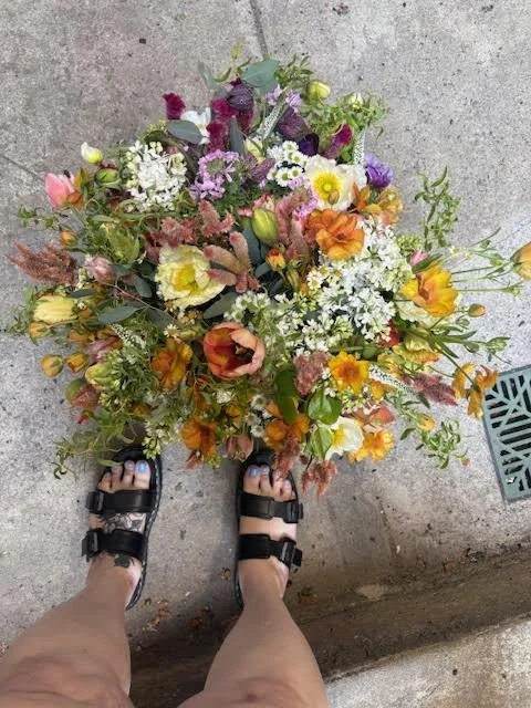 Top view of a person standing in sandals next to a large colorful bouquet of various flowers on a concrete surface.