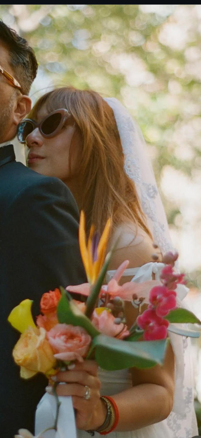 A man in a tuxedo and sunglasses stands in front of a woman with red hair, sunglasses, and a white veil, holding a colorful bouquet of flowers, all outdoors with blurred trees in the background.