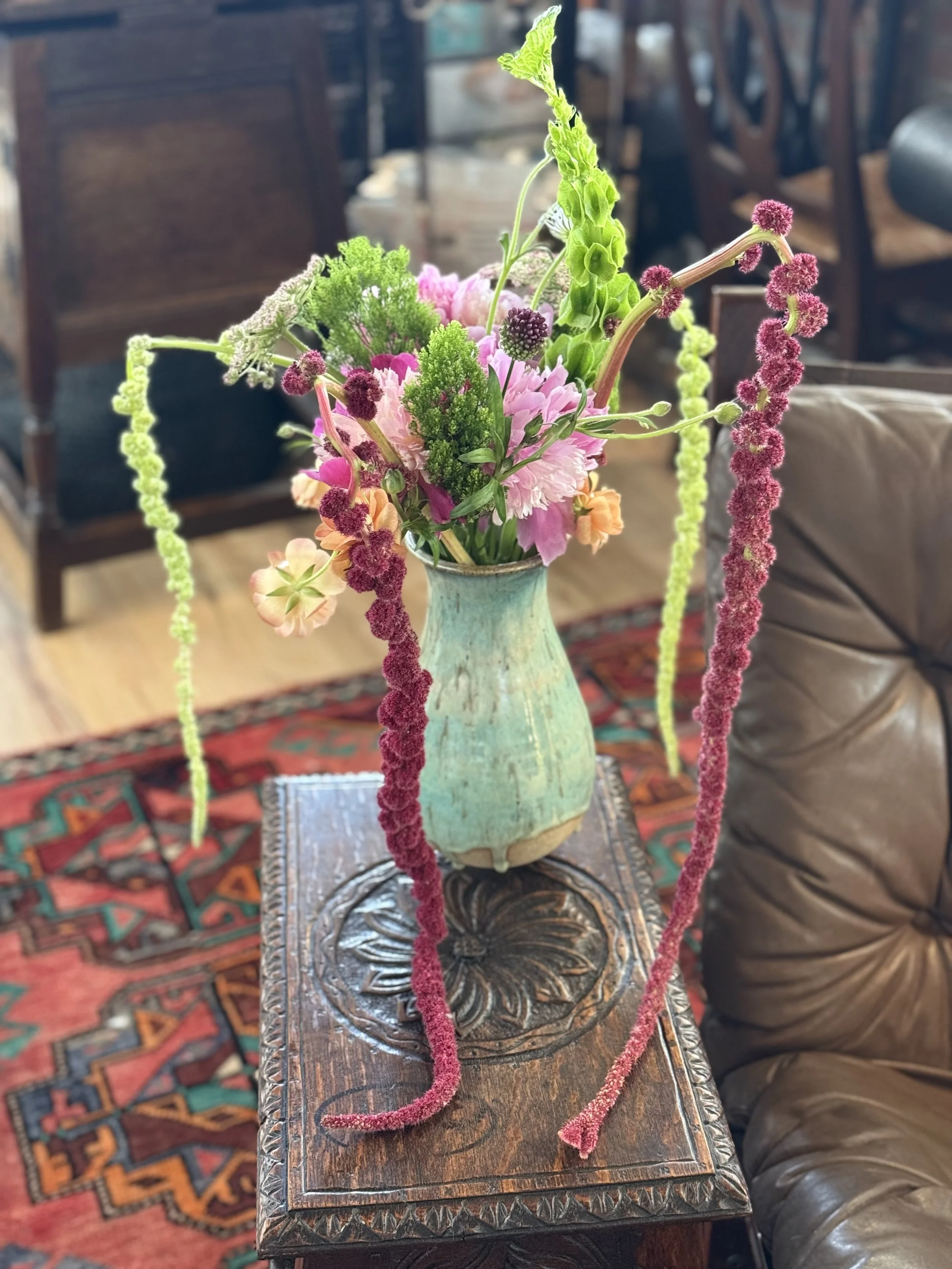 A green ceramic vase with a variety of colorful flowers, including pink, purple, and orange blooms, on a wooden table with carved floral designs. Three hanging pink and green yarn tassels drape from the vase.