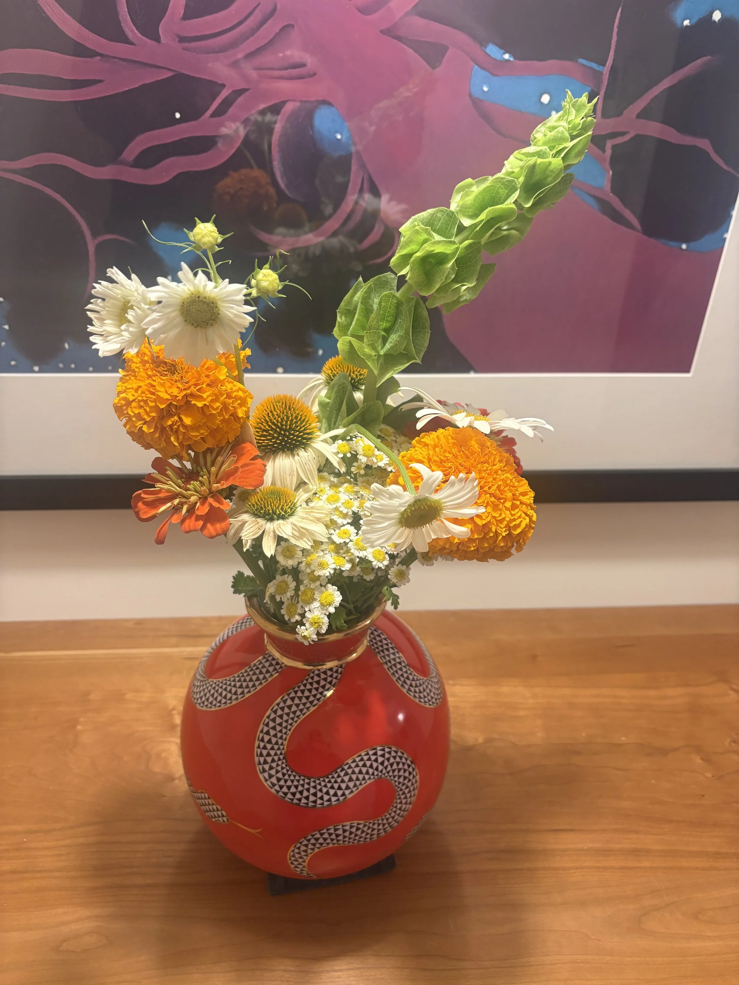 A colorful flower arrangement in a red vase with black and white snake patterns, featuring white daisies, orange marigolds, small white daisies, and green foliage, placed on a wooden surface.