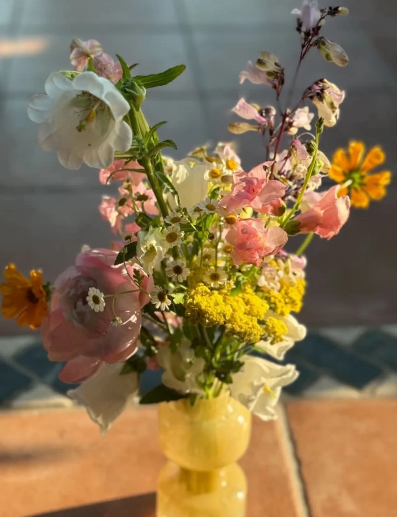 Colorful bouquet of mixed flowers in a yellow vase, placed on a tiled floor, with sunlight casting shadows.