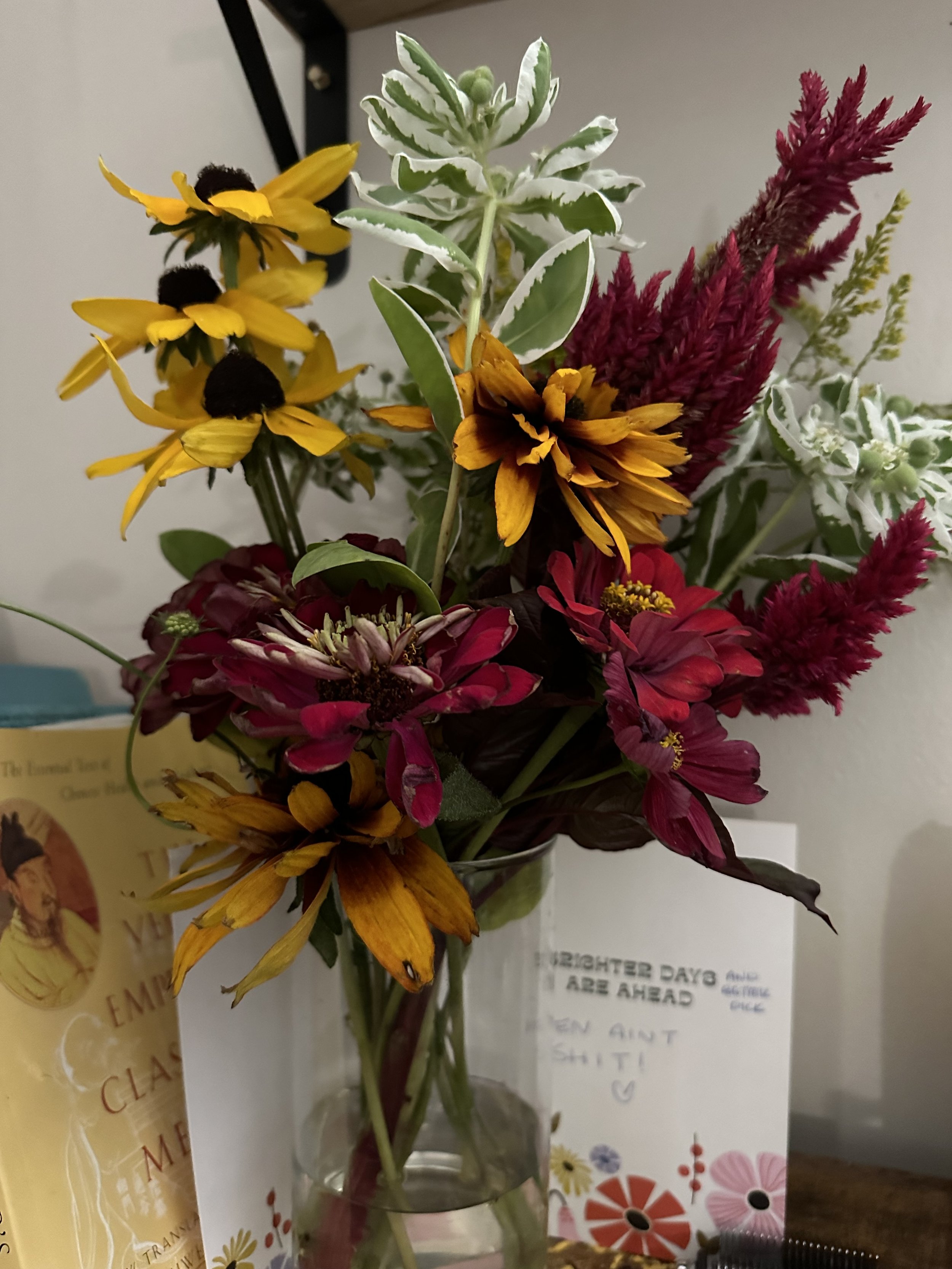 A bouquet of colorful flowers in a glass vase, including yellow daisies, red and pink zinnias, and burgundy astilbe, arranged against a neutral background.