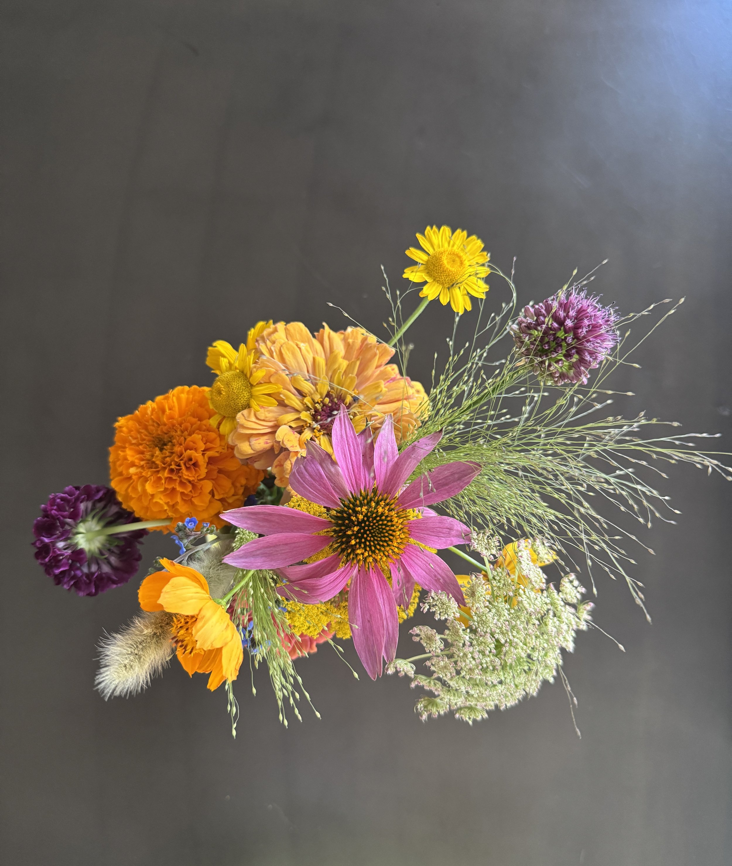 A colorful bouquet of various flowers, including a large pink flower, orange marigolds, yellow daisies, purple alliums, and white wildflowers, arranged on a dark surface.