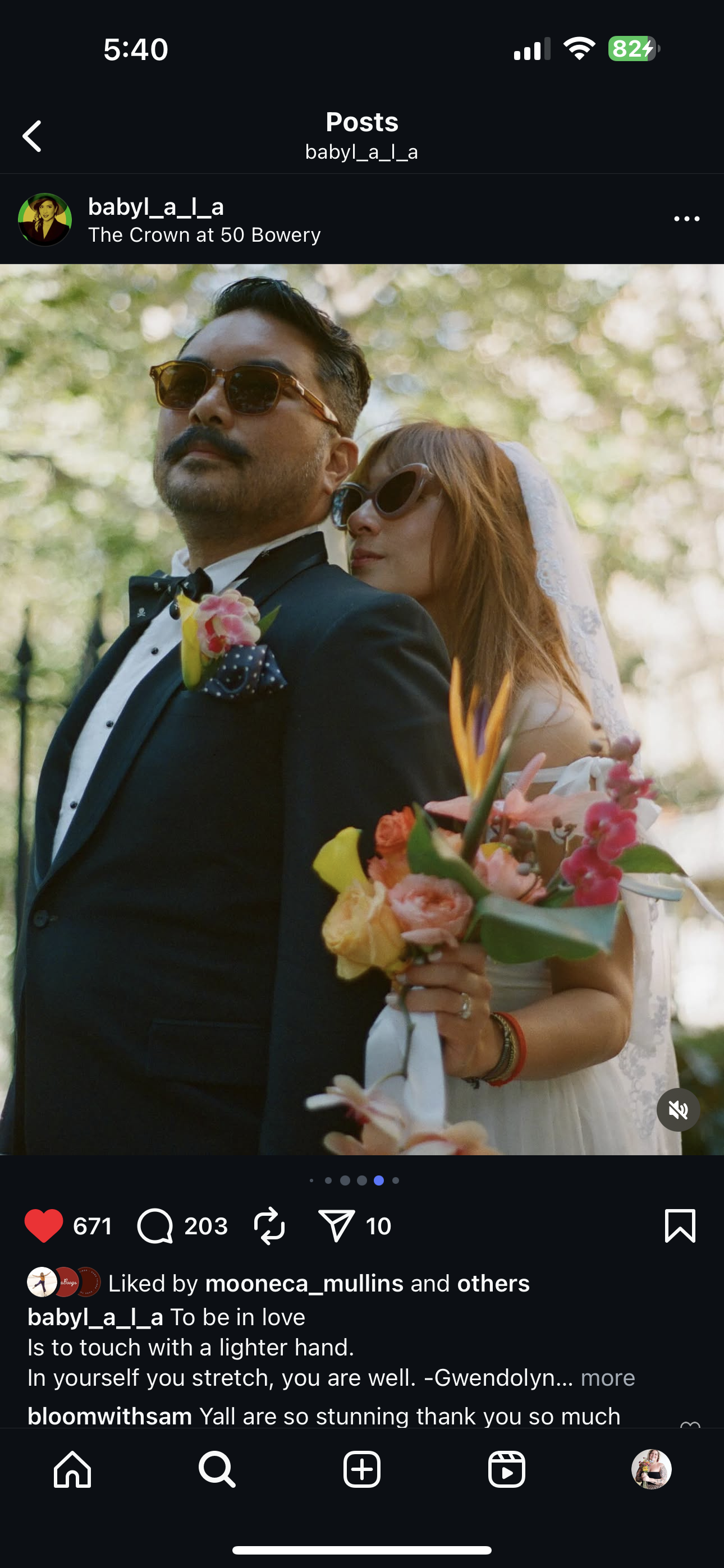 A man in a tuxedo and sunglasses stands in front of a woman with red hair, sunglasses, and a white veil, holding a colorful bouquet of flowers, all outdoors with blurred trees in the background.