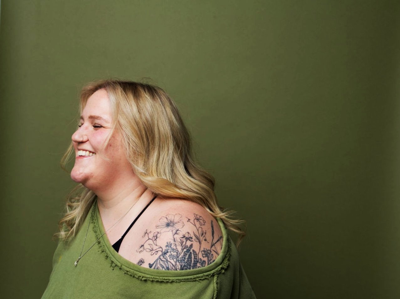 Side view of a smiling woman with blonde hair, wearing a green top and a necklace, showing a floral tattoo on her shoulder.