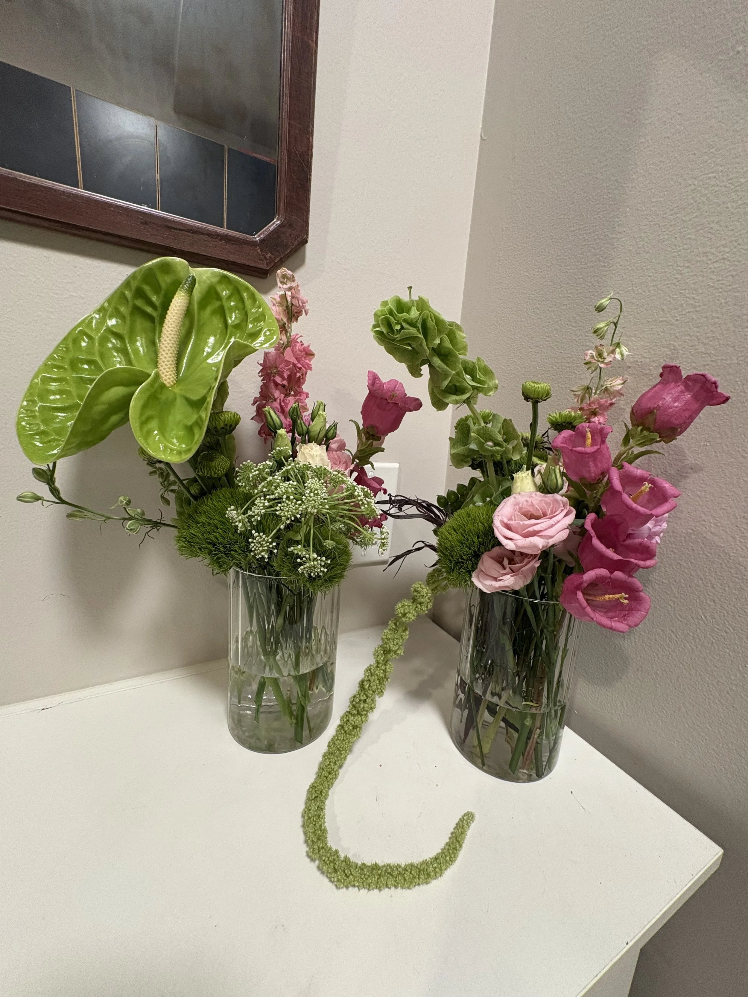 Two glass vases with pink, green, and white flowers on a white surface, with a beige wall and mirror in the background.