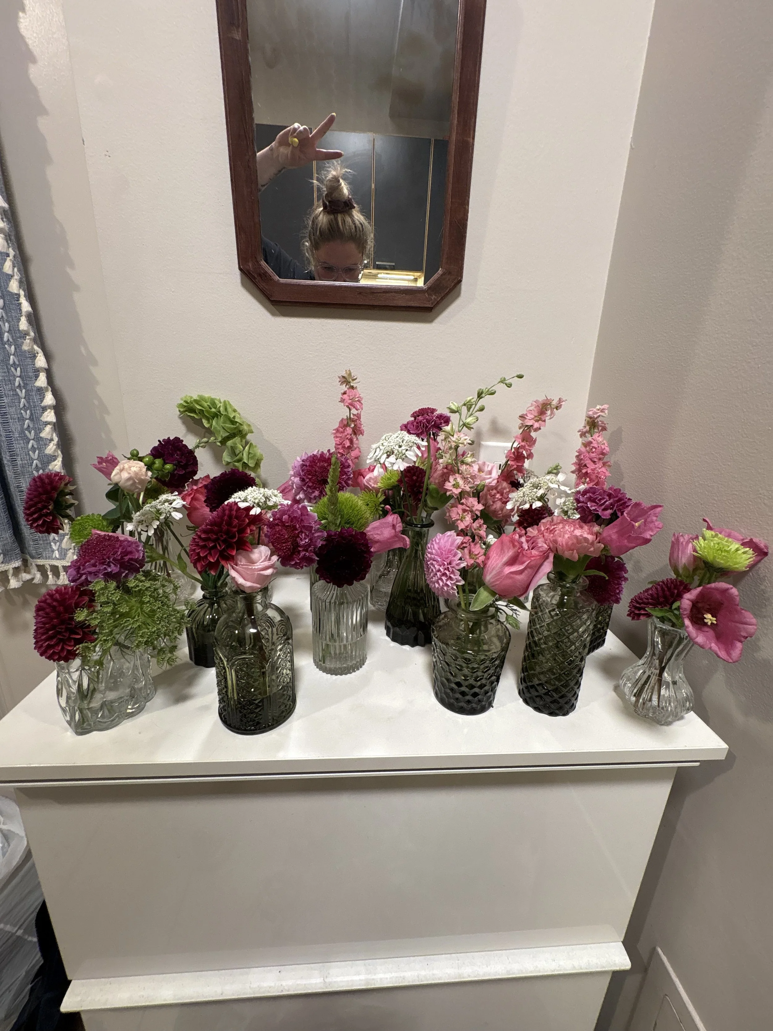 A white dresser topped with multiple glass vases filled with pink and purple flowers. A mirror above the dresser shows a woman making a peace sign, with her hair in a bun and wearing glasses.