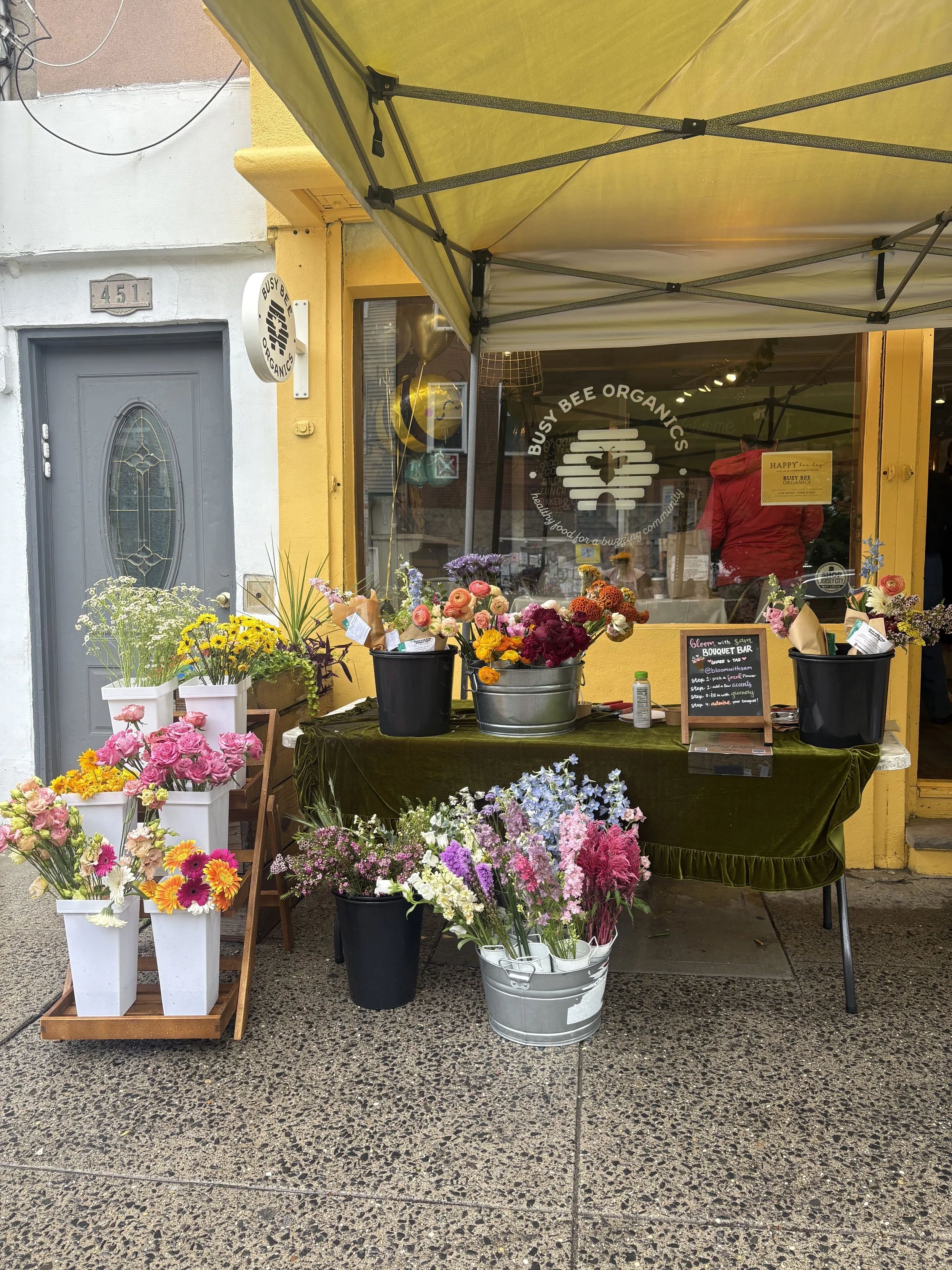Street flower stand with various colorful flowers in buckets and vases outside a yellow storefront, with a sign for Busy Bee Organics visible through the window.