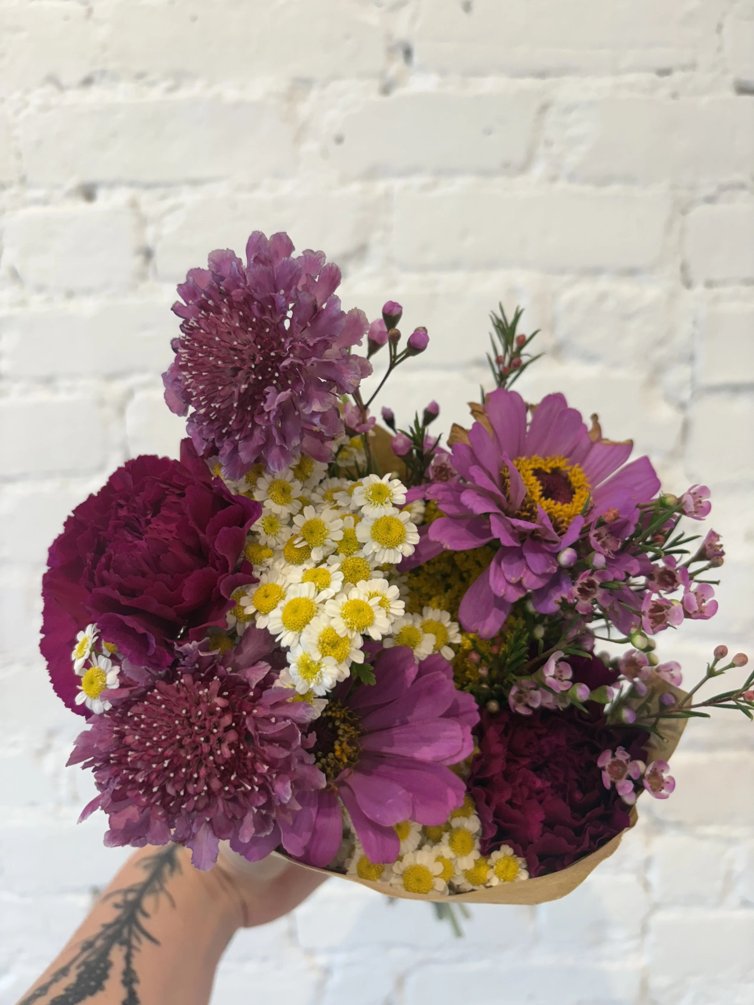 A colorful bouquet of various pink, purple, and white flowers held against a white brick wall.