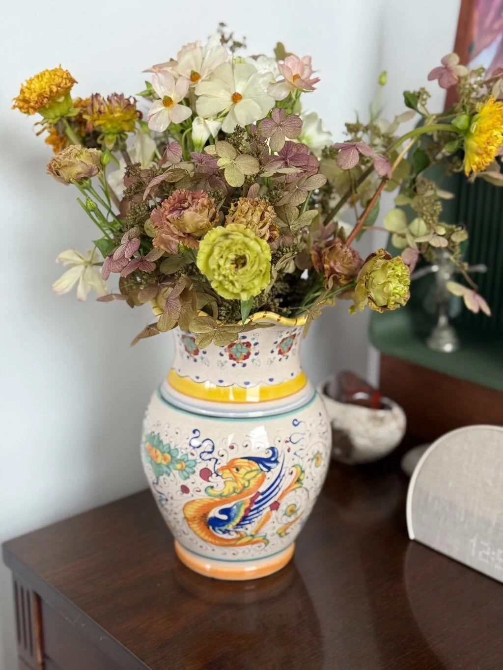 A colorful vase with floral patterns containing a bouquet of dried flowers on a wooden table.